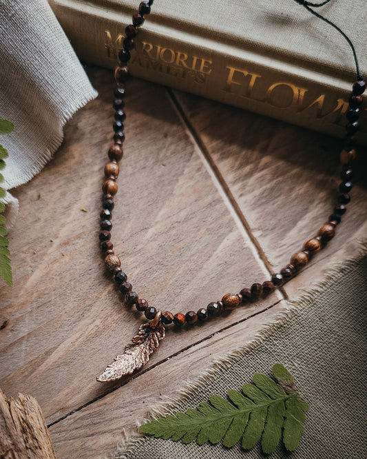 Necklace with red tiger's eye and wooden beads and a copper electroformed fern charm on a wooden surface with a book and fern leaf.