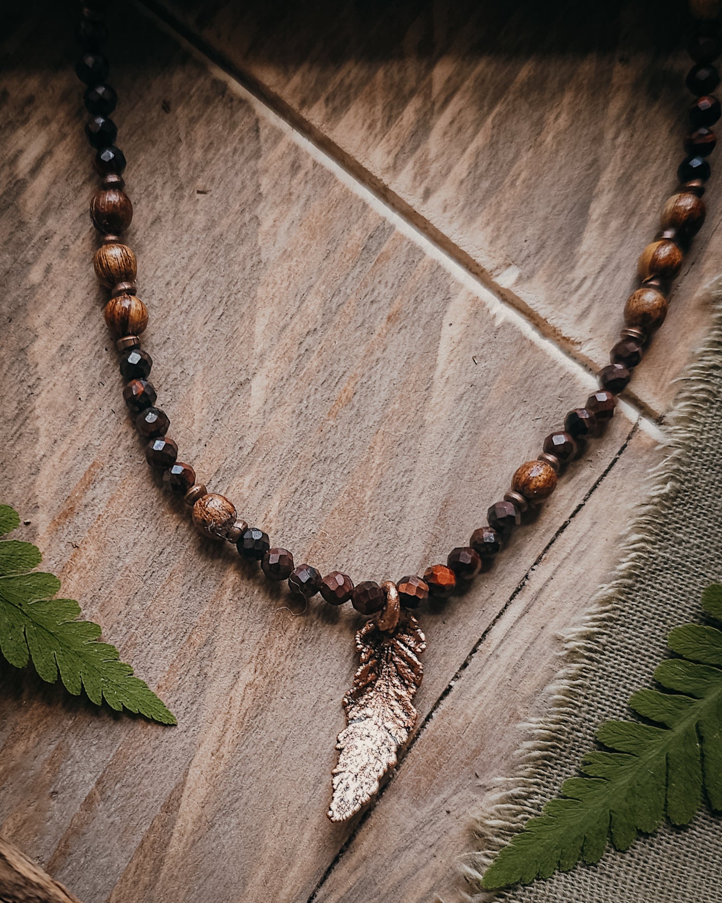 Necklace with red tiger's eye and wooden beads and a copper electroformed fern charm on a wooden surface with a book and fern leaf.