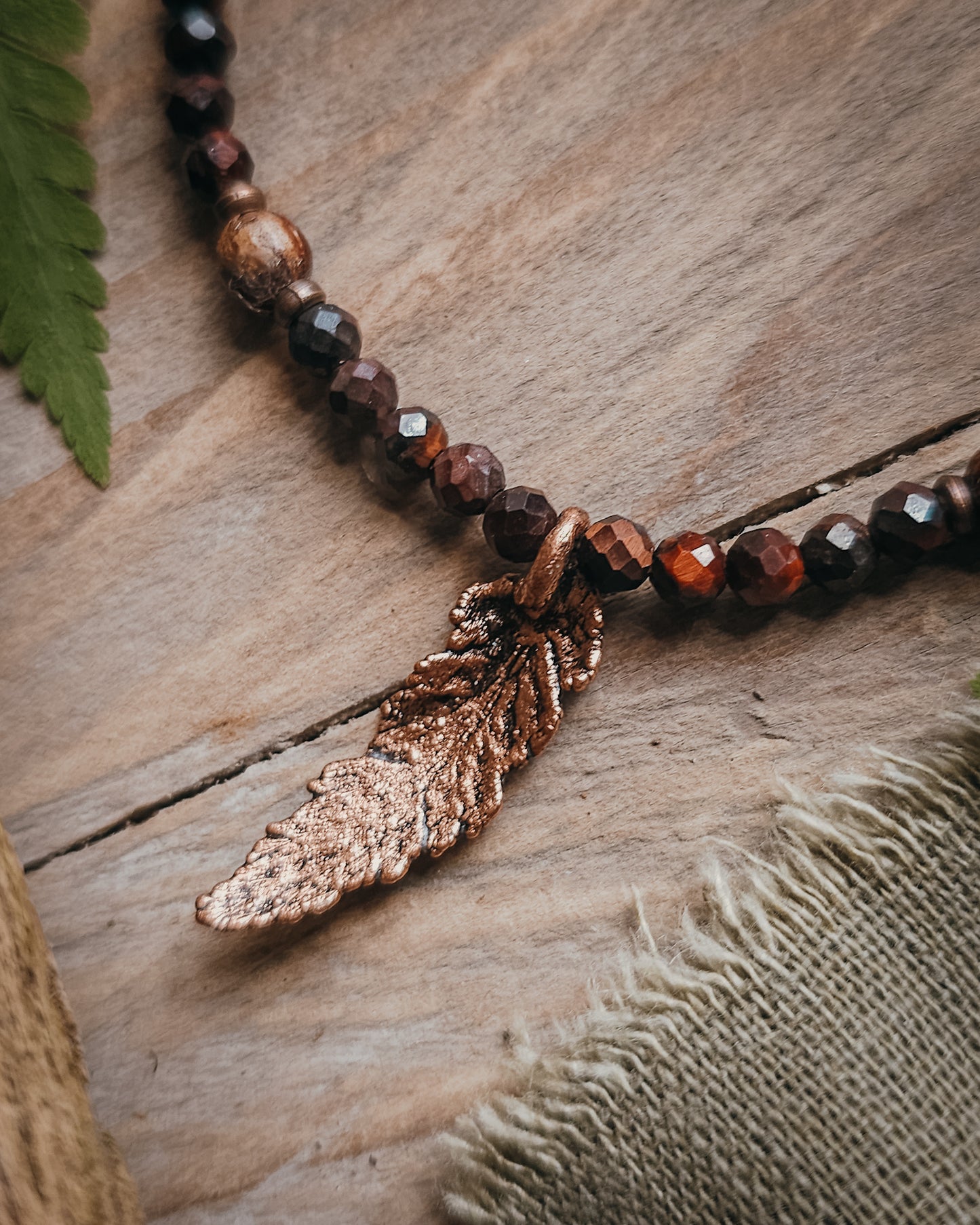 Necklace with red tiger's eye and wooden beads and a copper electroformed fern charm on a wooden surface with a book and fern leaf.