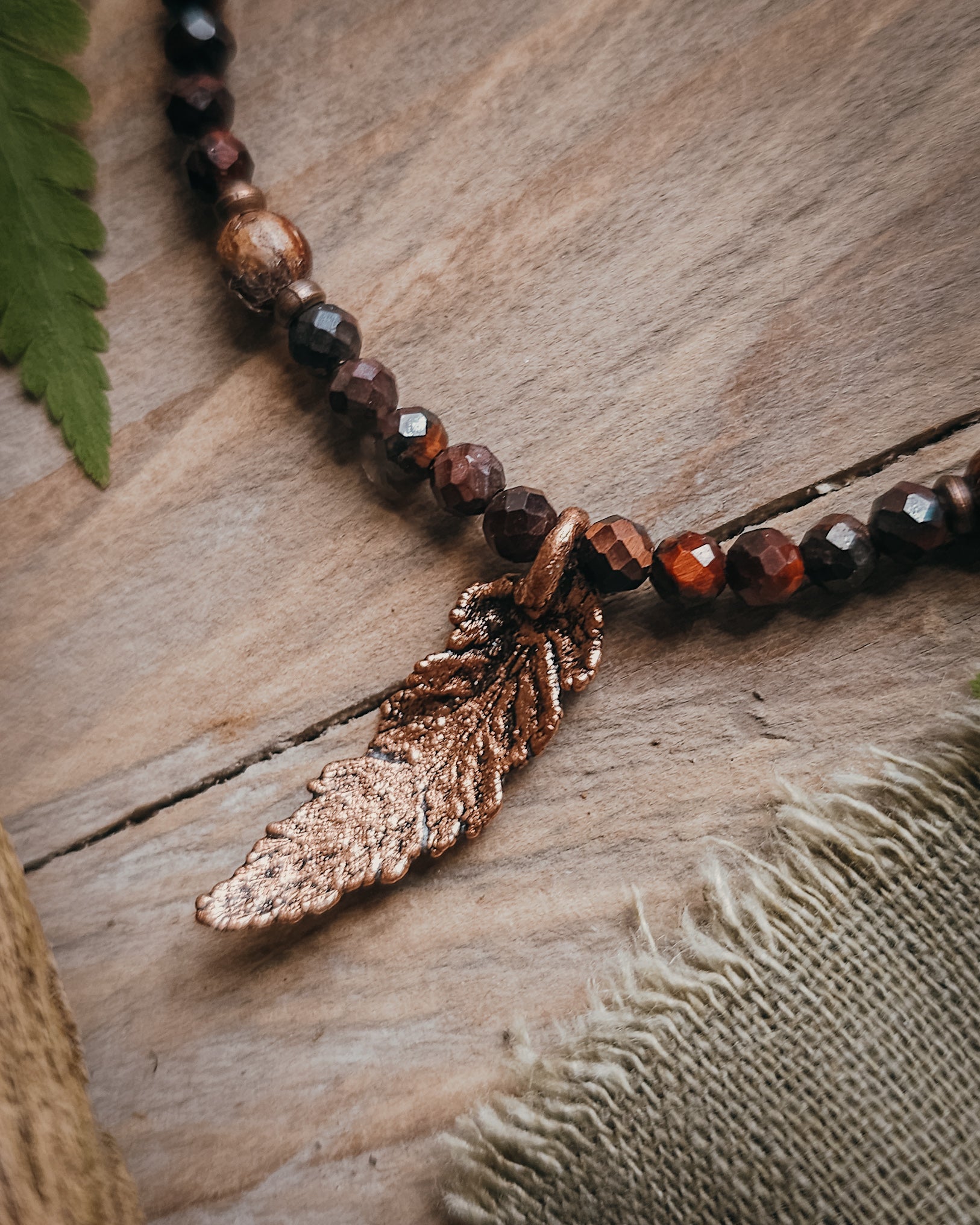 Necklace with red tiger's eye and wooden beads and a copper electroformed fern charm on a wooden surface with a book and fern leaf.