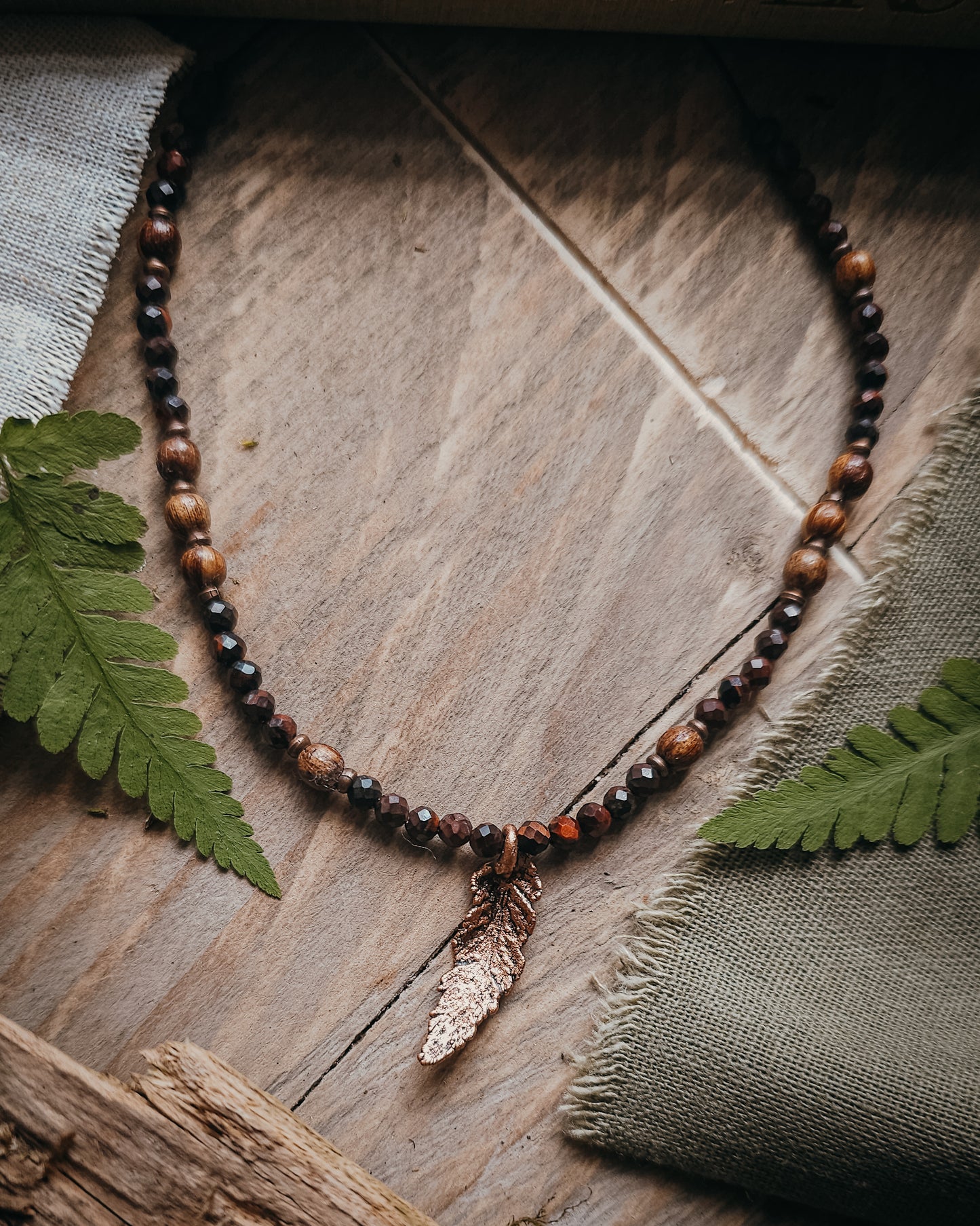 Necklace with red tiger's eye and wooden beads and a copper electroformed fern charm on a wooden surface with a book and fern leaf.
