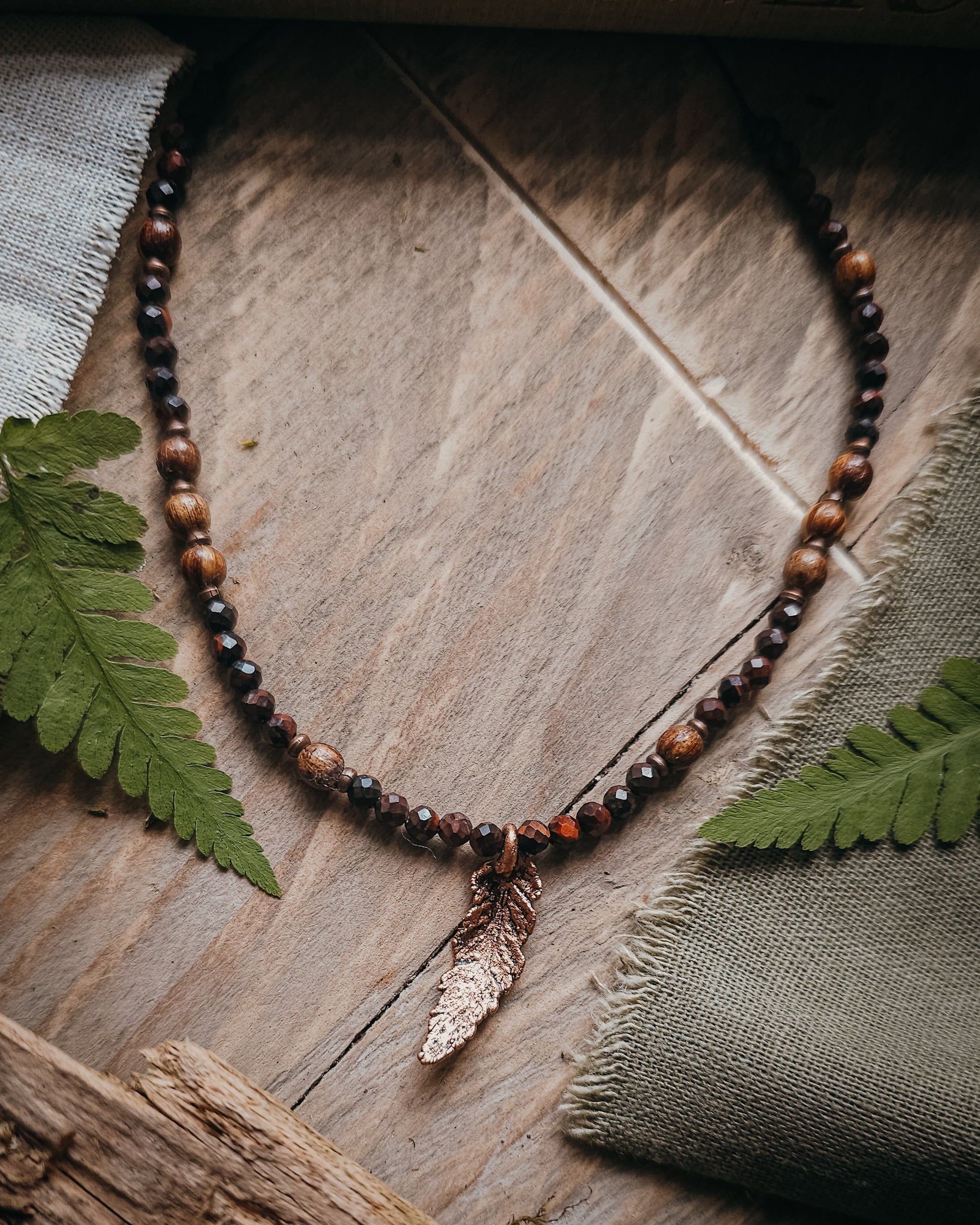 Necklace with red tiger's eye and wooden beads and a copper electroformed fern charm on a wooden surface with a book and fern leaf.