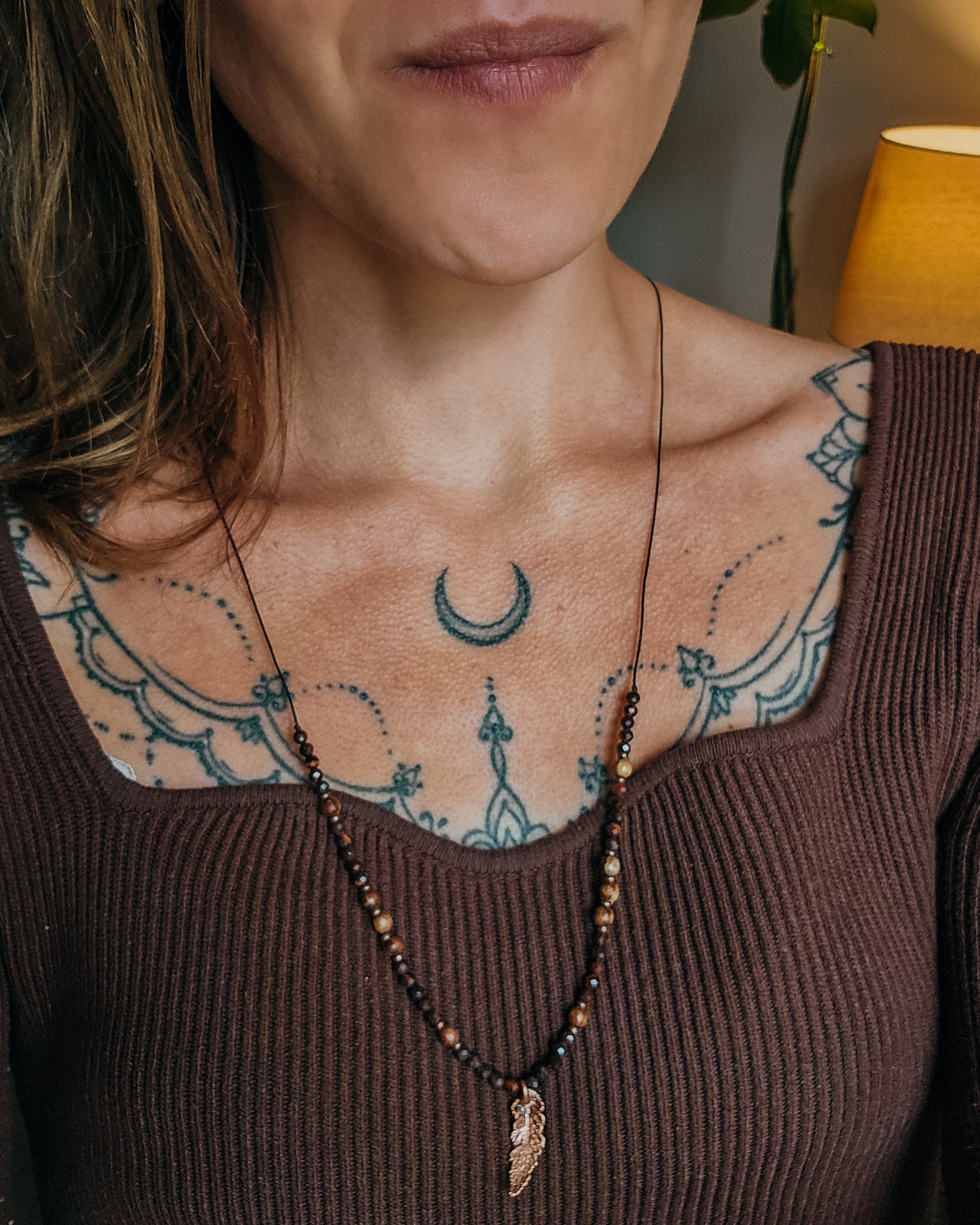 Close-up of a woman with tattoos on their shoulder and neck, wearing Necklace with red tiger's eye and wooden beads and a copper electroformed fern charm on a wooden surface with a book and fern leaf.