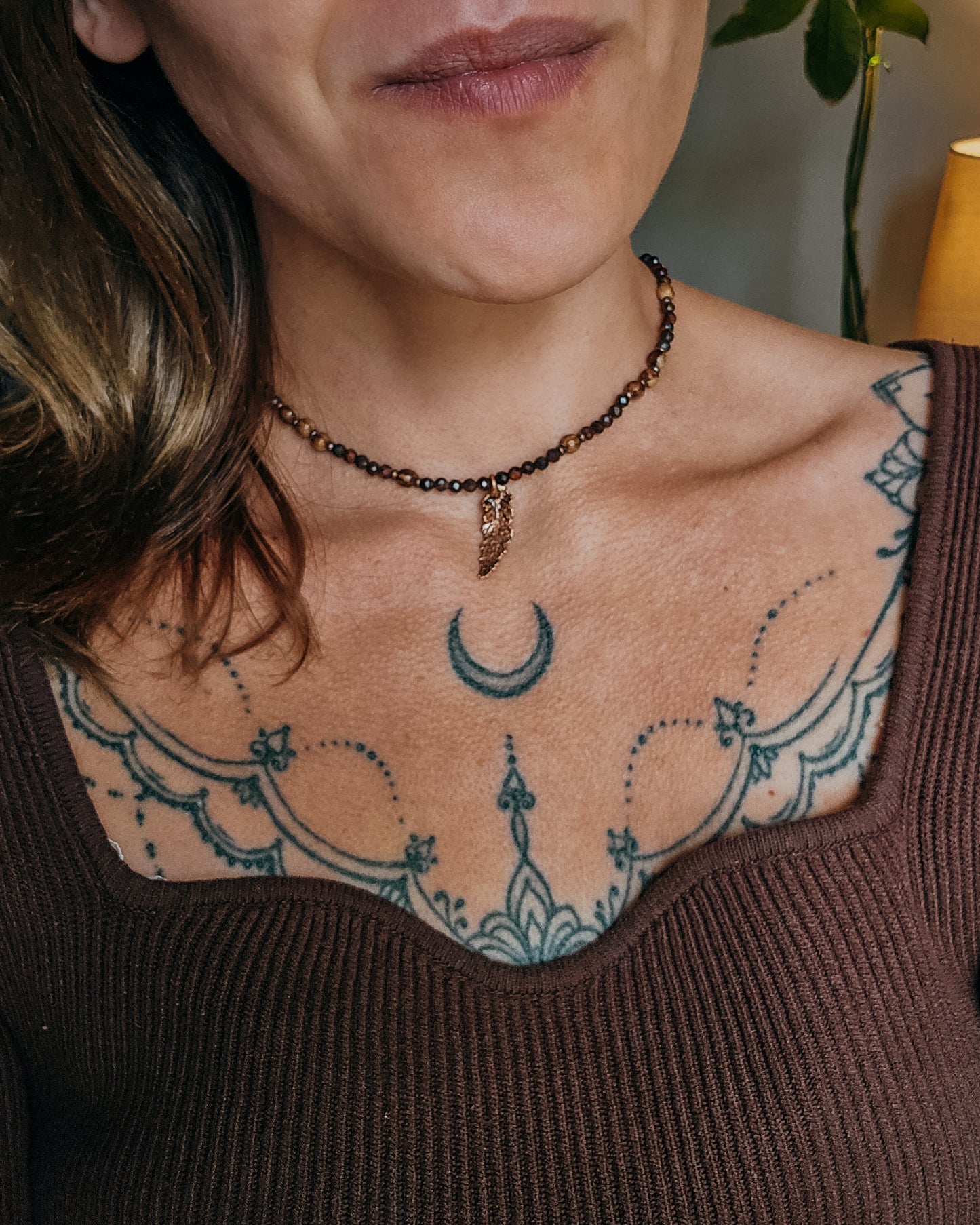 Close-up of a woman with tattoos on their shoulder and neck, wearing Necklace with red tiger's eye and wooden beads and a copper electroformed fern charm on a wooden surface with a book and fern leaf.