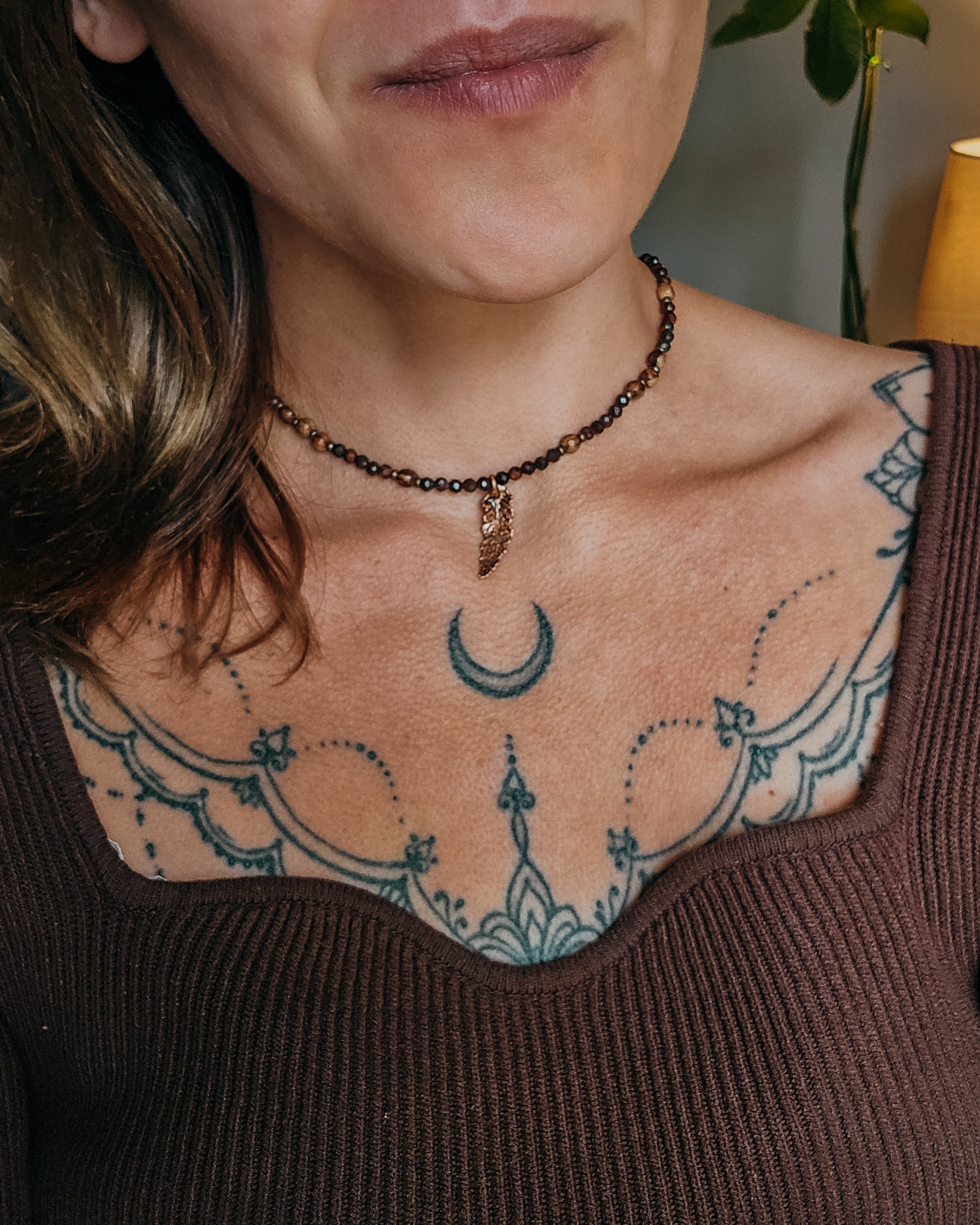 Close-up of a woman with tattoos on their shoulder and neck, wearing Necklace with red tiger's eye and wooden beads and a copper electroformed fern charm on a wooden surface with a book and fern leaf.