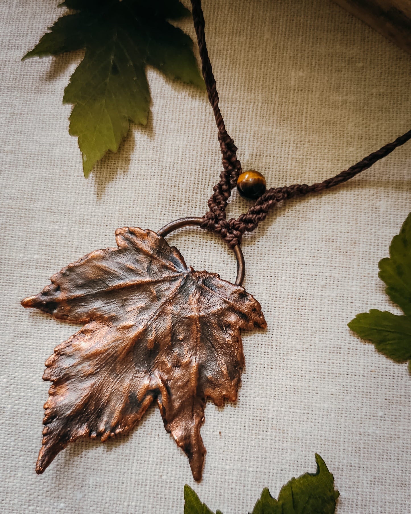 Handmade Copper electroformed Necklace with maple leaf pendant, macrame and Tiger's Eye bead on a textured surface with leaves
