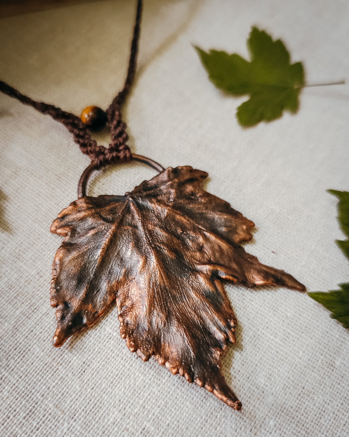 Handmade Copper electroformed Necklace with maple leaf pendant, macrame and Tiger's Eye bead on a textured surface with leaves