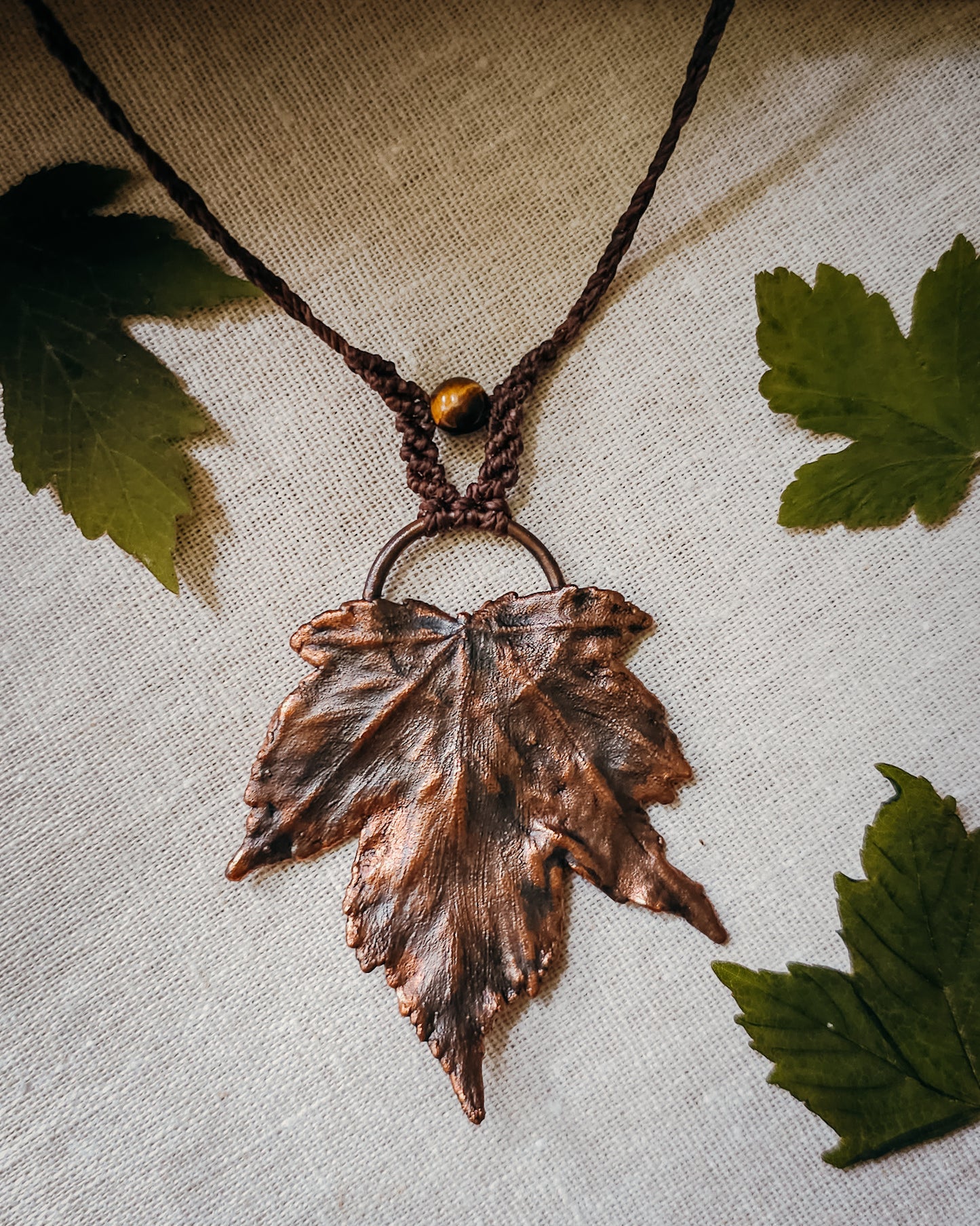 Handmade Copper electroformed Necklace with maple leaf pendant, macrame and Tiger's Eye bead on a textured surface with leaves