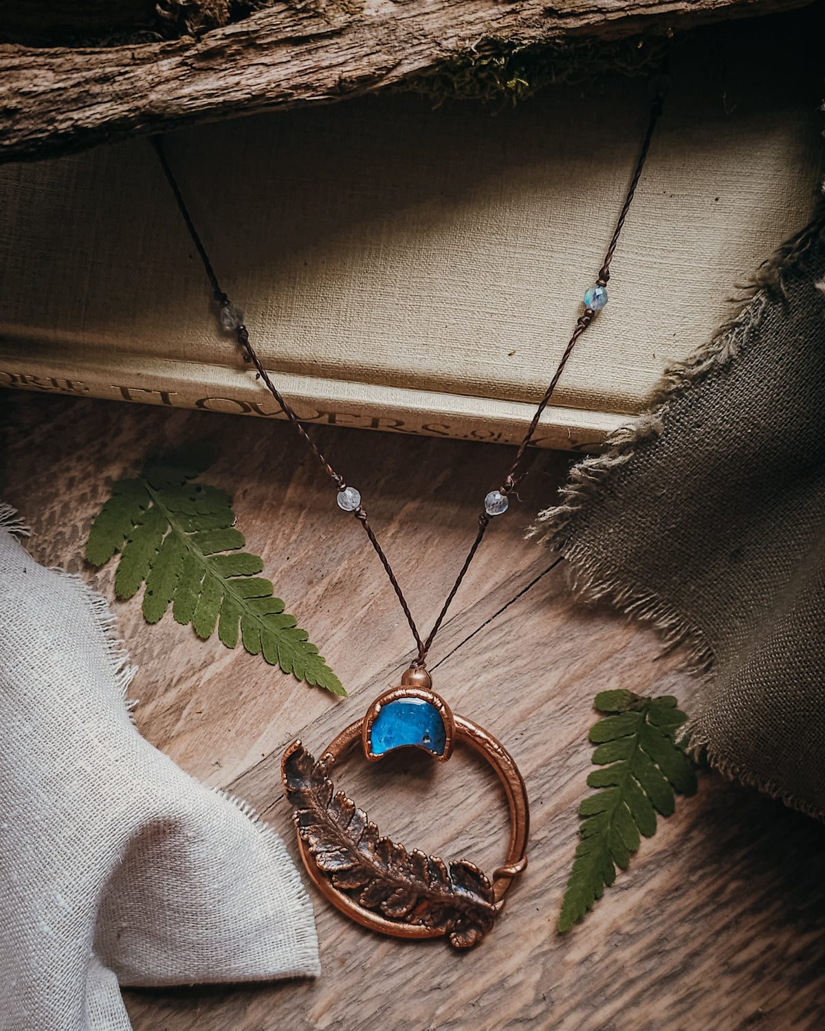 Copper electroformed fern pendant with a Labradorite crescent moon set within a circular copper frame, suspended from an adjustable cord with Labradorite and brass accent beads, handcrafted from a real fern gathered in Scotland and photographed on a rustic woodland-inspired background.