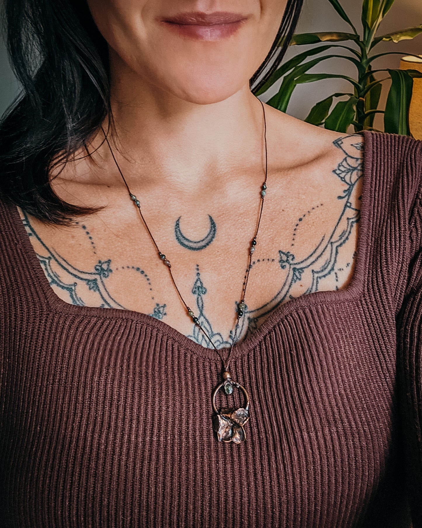 Handcrafted copper Primrose flower necklace worn on an adjustable cord, featuring a Green Tourmaline stone, green and pink Tourmaline beads, and intricate details. The real Scottish Primrose preserved in copper is shown worn against the neckline for scale.