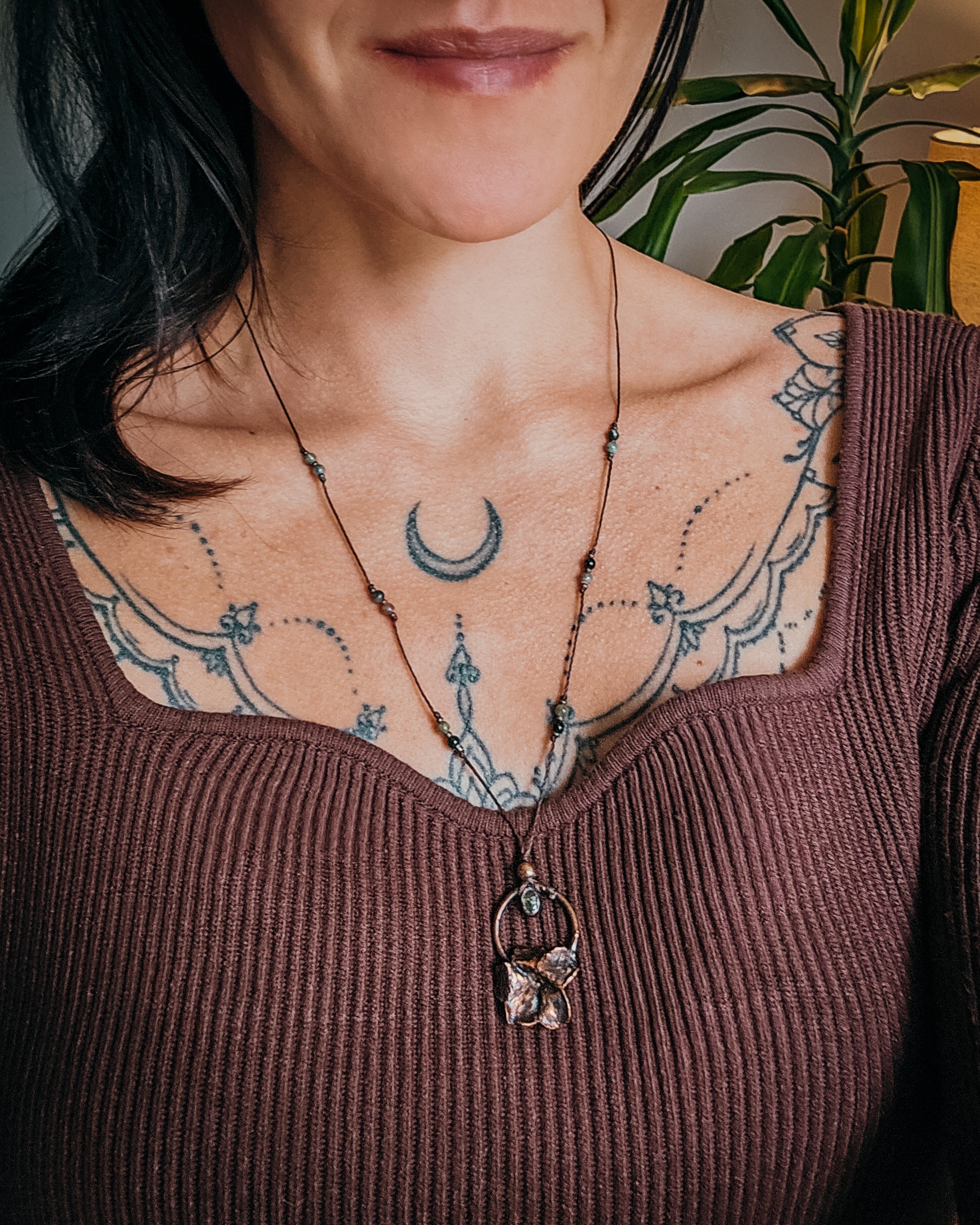 Handcrafted copper Primrose flower necklace worn on an adjustable cord, featuring a Green Tourmaline stone, green and pink Tourmaline beads, and intricate details. The real Scottish Primrose preserved in copper is shown worn against the neckline for scale.