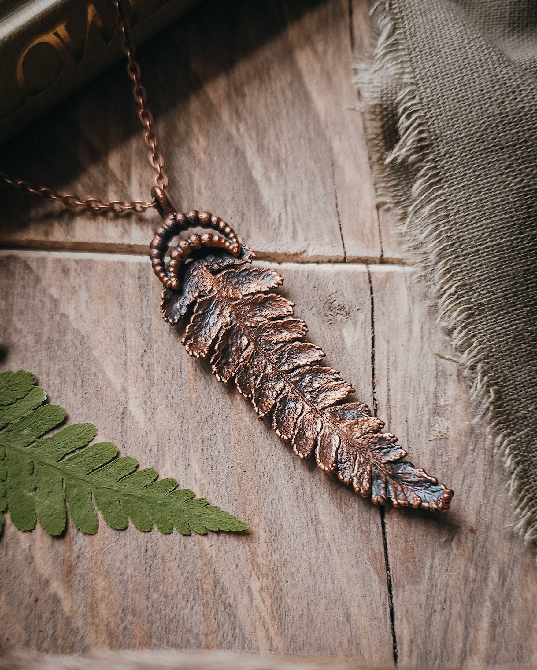 Copper electroformed fern pendant with crescent moon detail on a copper-coloured stainless steel chain, handcrafted from a real fern gathered in Scotland and photographed with fresh fern fronds on a rustic woodland-inspired background.