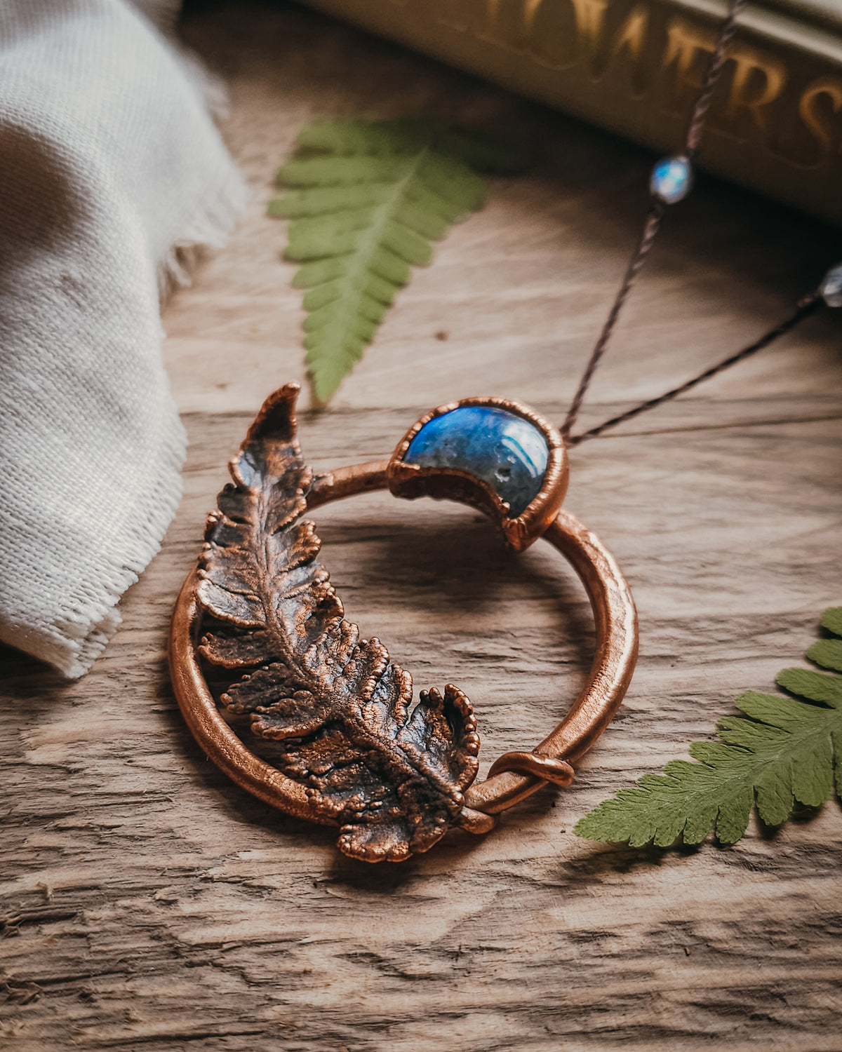 Copper electroformed fern pendant with a Labradorite crescent moon set within a circular copper frame, suspended from an adjustable cord with Labradorite and brass accent beads, handcrafted from a real fern gathered in Scotland and photographed on a rustic woodland-inspired background.