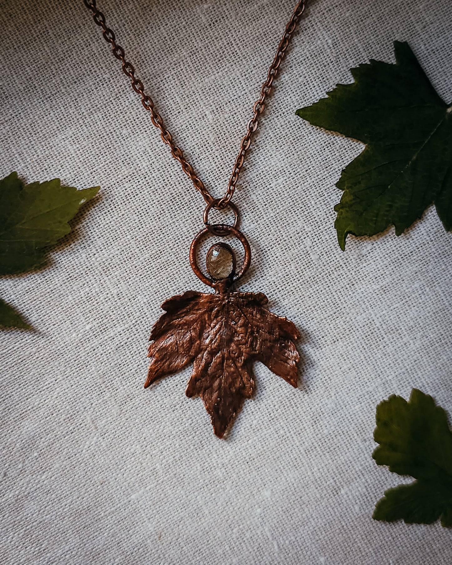 Copper electroformed Necklace with maple leaf pendant and golden rutilated Quartz crystal on a textured surface with leaves