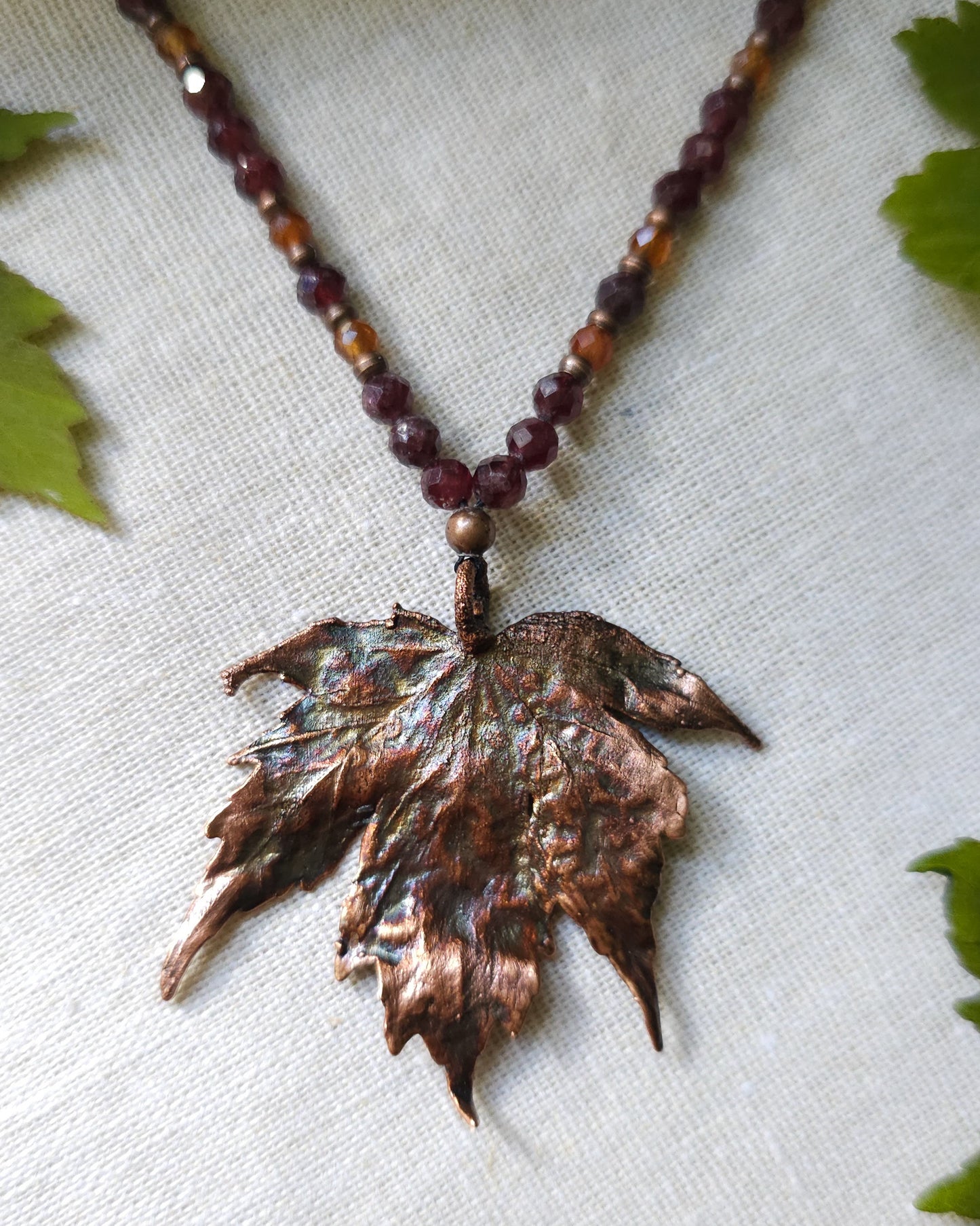 Handmade Copper electroformed Necklace with maple leaf pendant and Garnet crystal beads on a textured surface with leaves