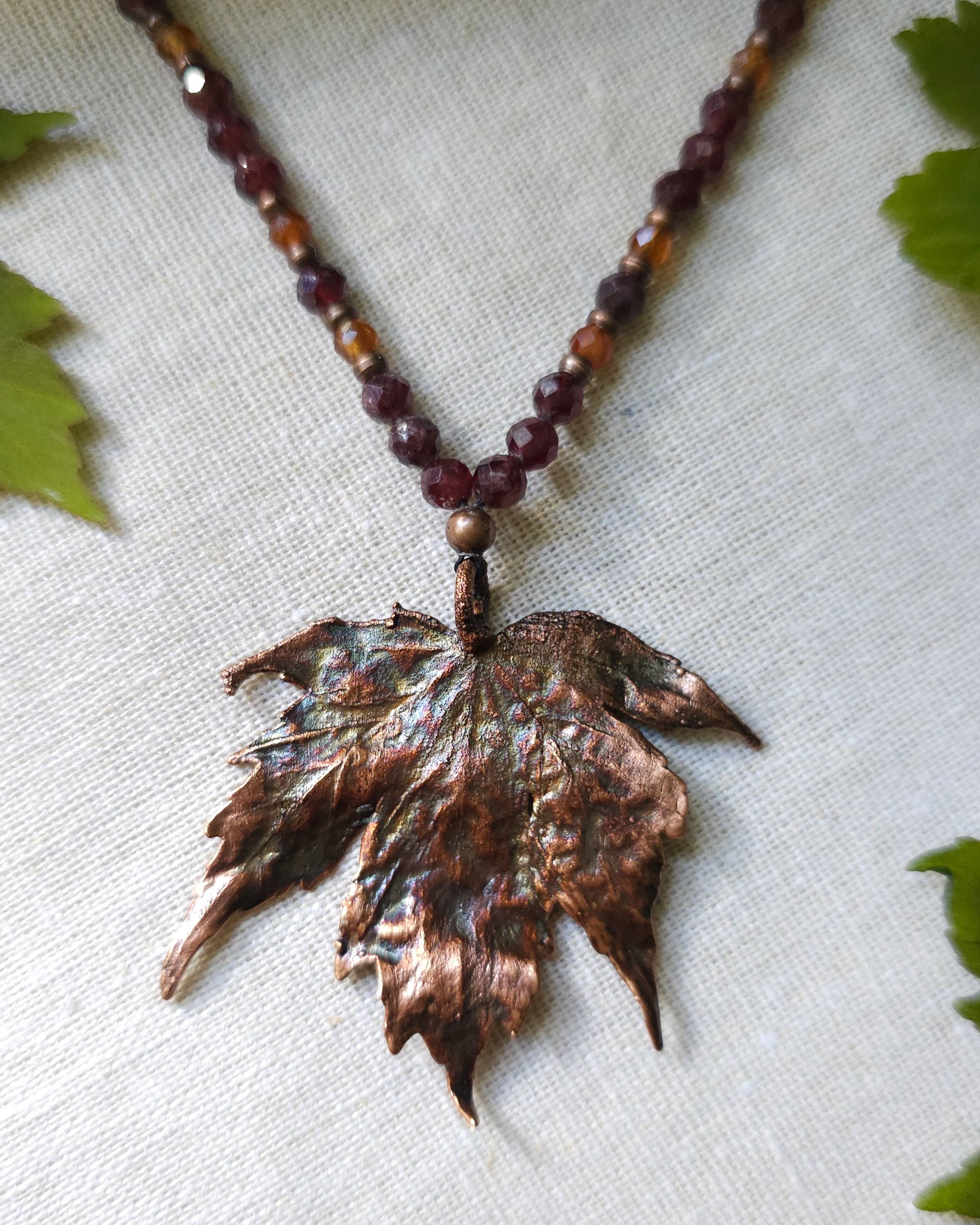 Handmade Copper electroformed Necklace with maple leaf pendant and Garnet crystal beads on a textured surface with leaves