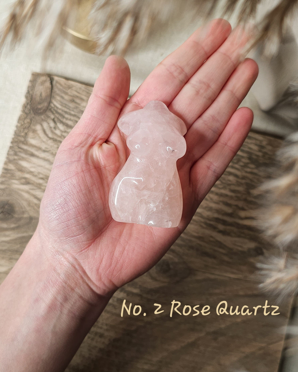 A hand holding a small carved pale pink rose quartz female body figurine, with a wood surface and soft neutral background behind it. 