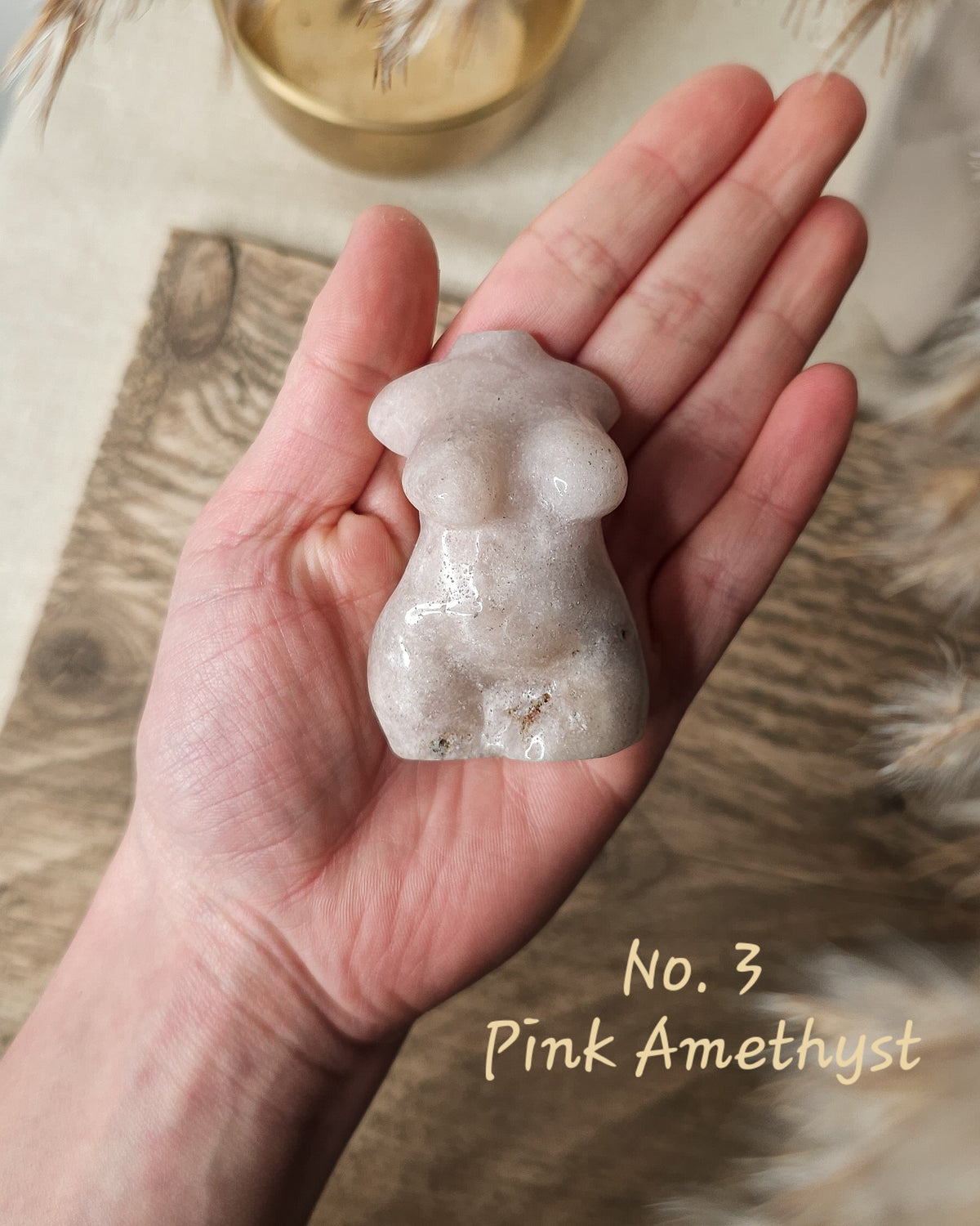 A hand holds a small polished pink amethyst crystal figurine shaped like a stylized torso. The crystal has a translucent pale pink tone with natural inclusions.