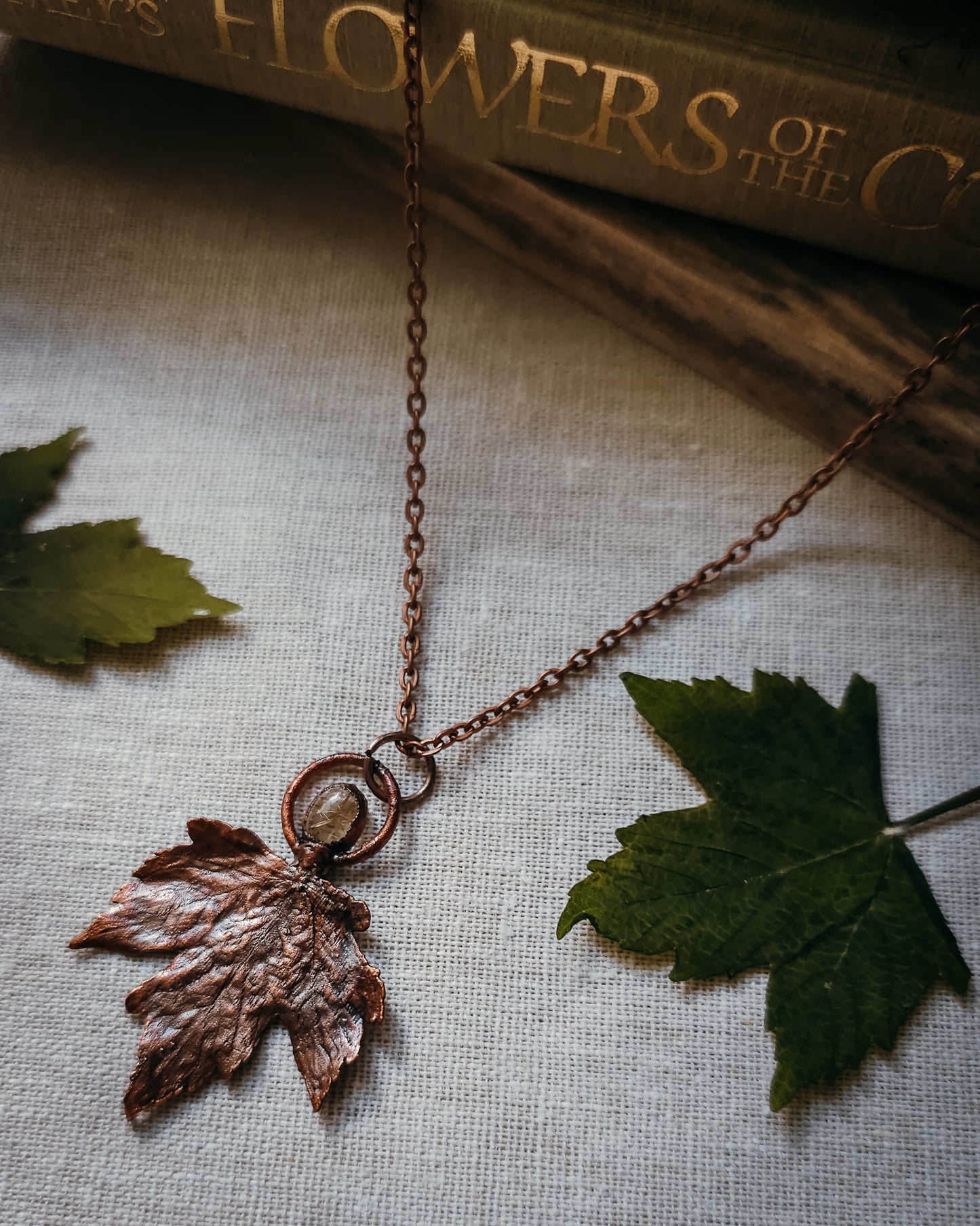 Copper electroformed Necklace with maple leaf pendant and golden rutilated Quartz crystal on a textured surface with leaves