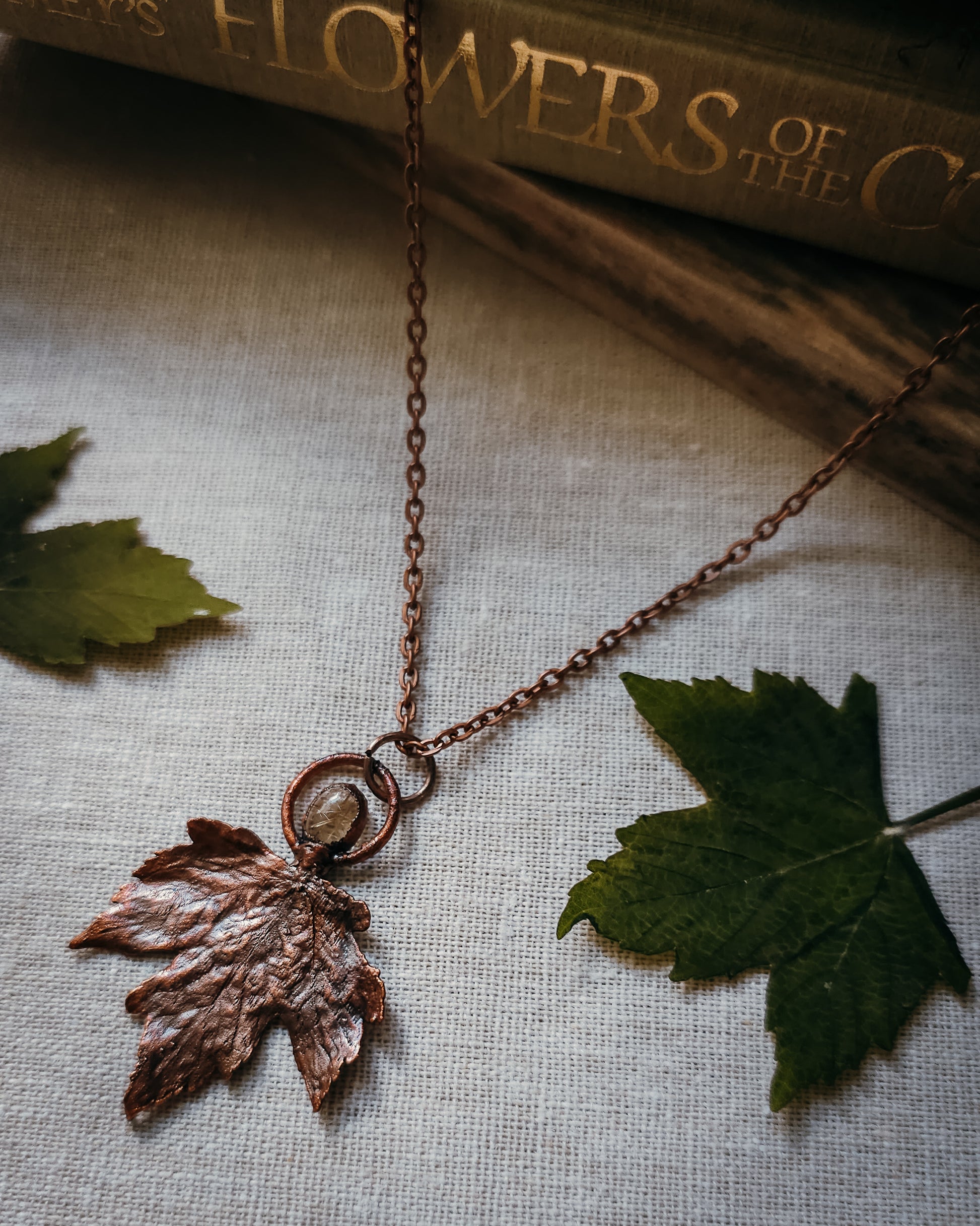 Copper electroformed Necklace with maple leaf pendant and golden rutilated Quartz crystal on a textured surface with leaves