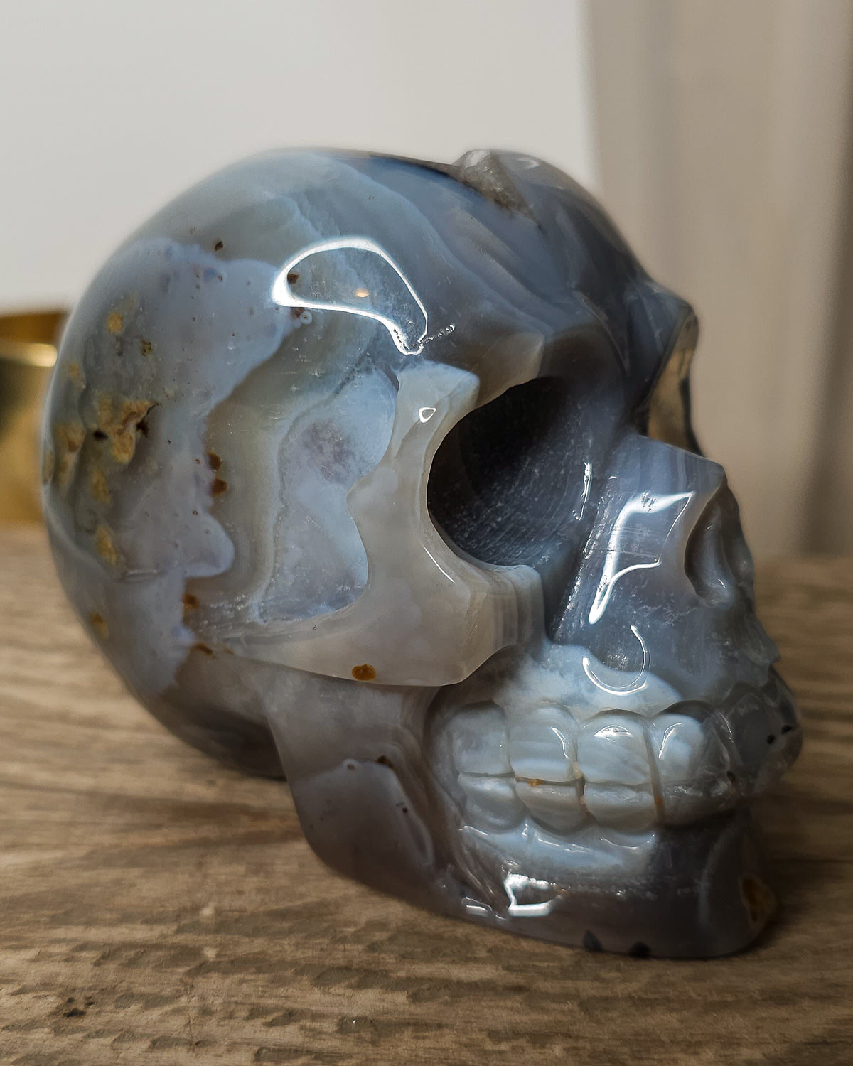 Blue Agate skull carving with layered pale blue and grey bands, a natural brown inclusion along the side and a sparkling druzy crystal cavity at the top.