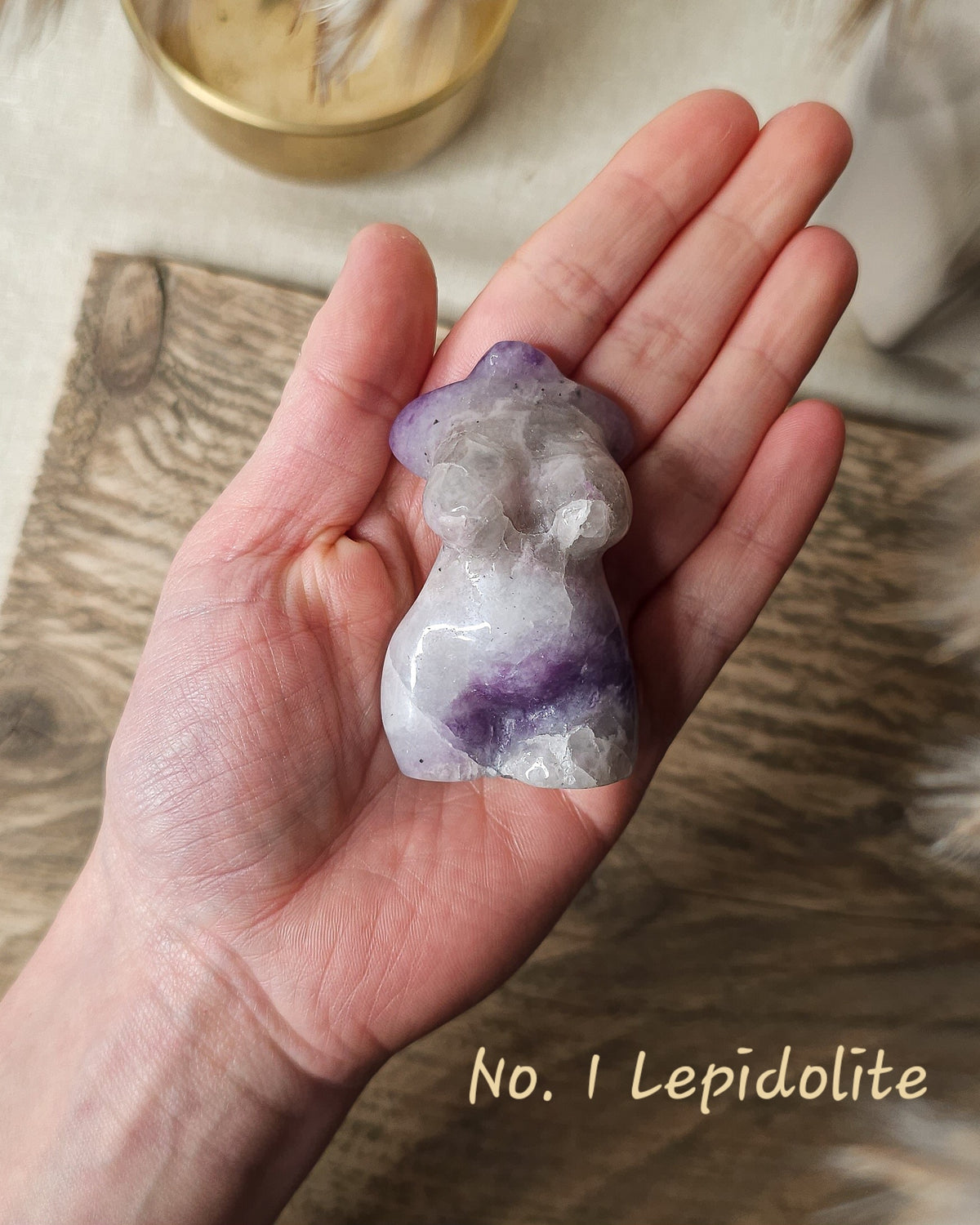 A hand holding a polished lepidolite crystal figurine shaped like a small human figure. The crystal shows natural purple and white mottling with a rough, raw-texture surface.