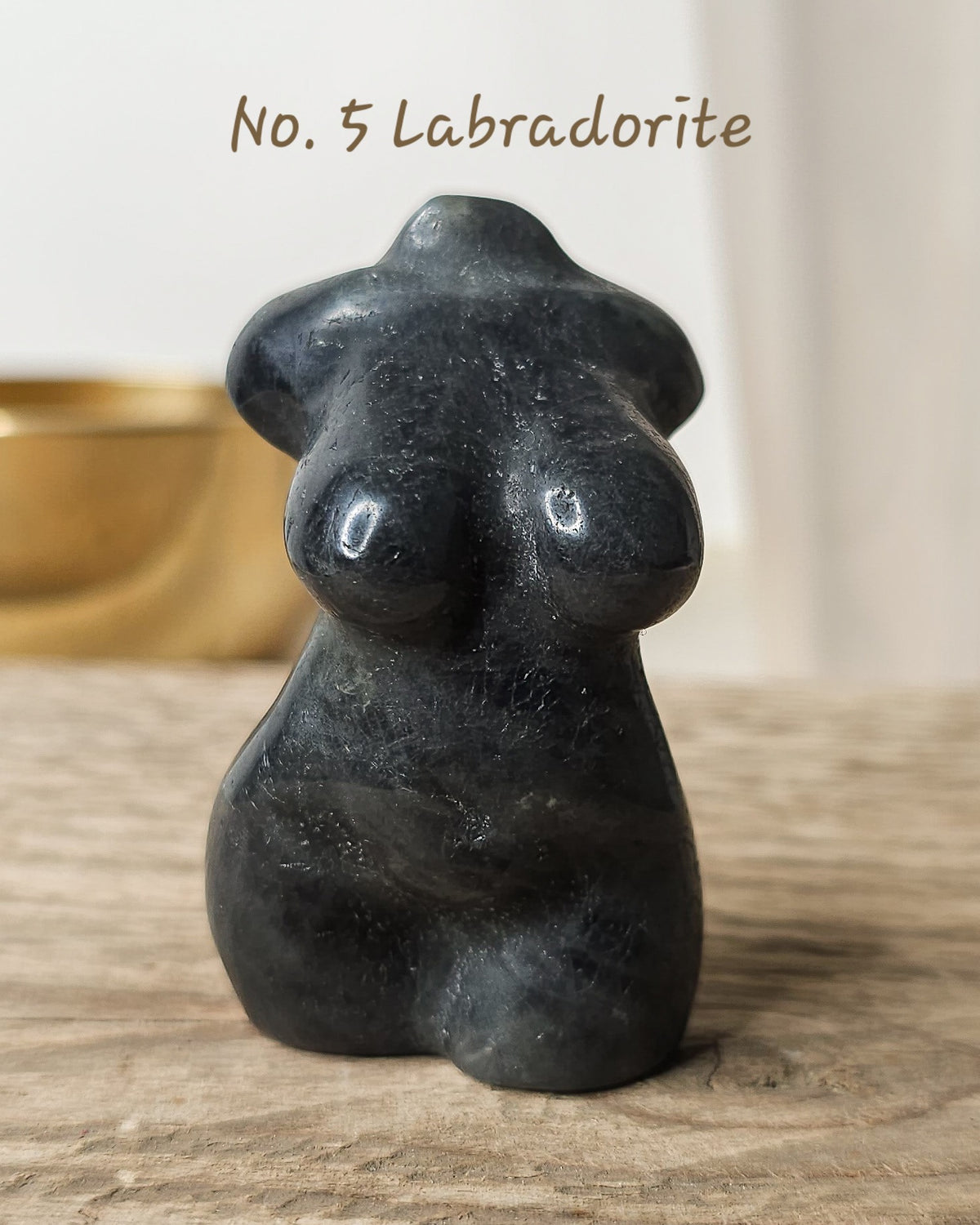 A small black labradorite stone sculpture shaped like a stylized female torso, standing on a rustic wooden surface with a softly blurred warm-toned background. 