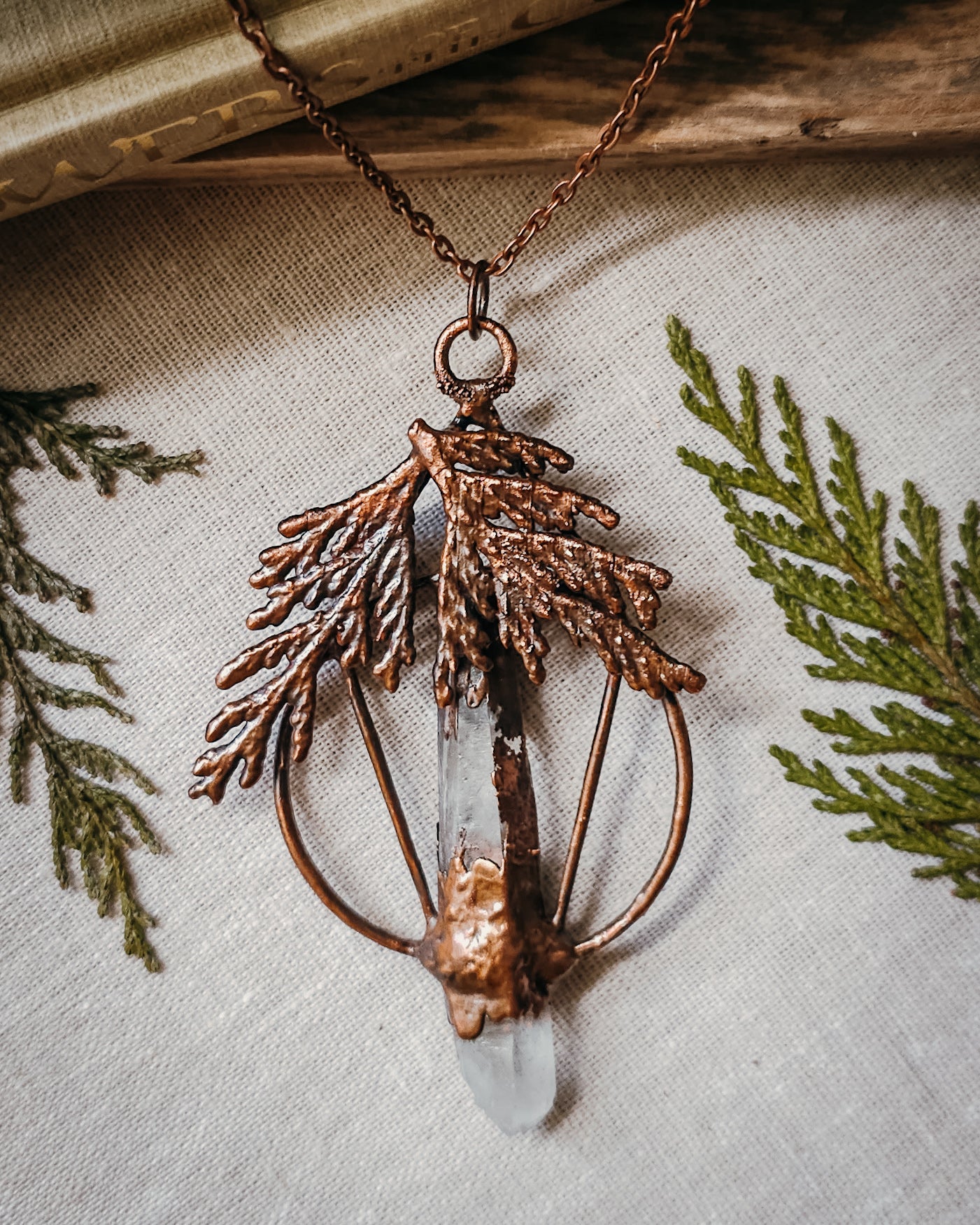Handmade Copper electroformed Necklace with a cypress and clear Quartz crystal pendant on a textured fabric background