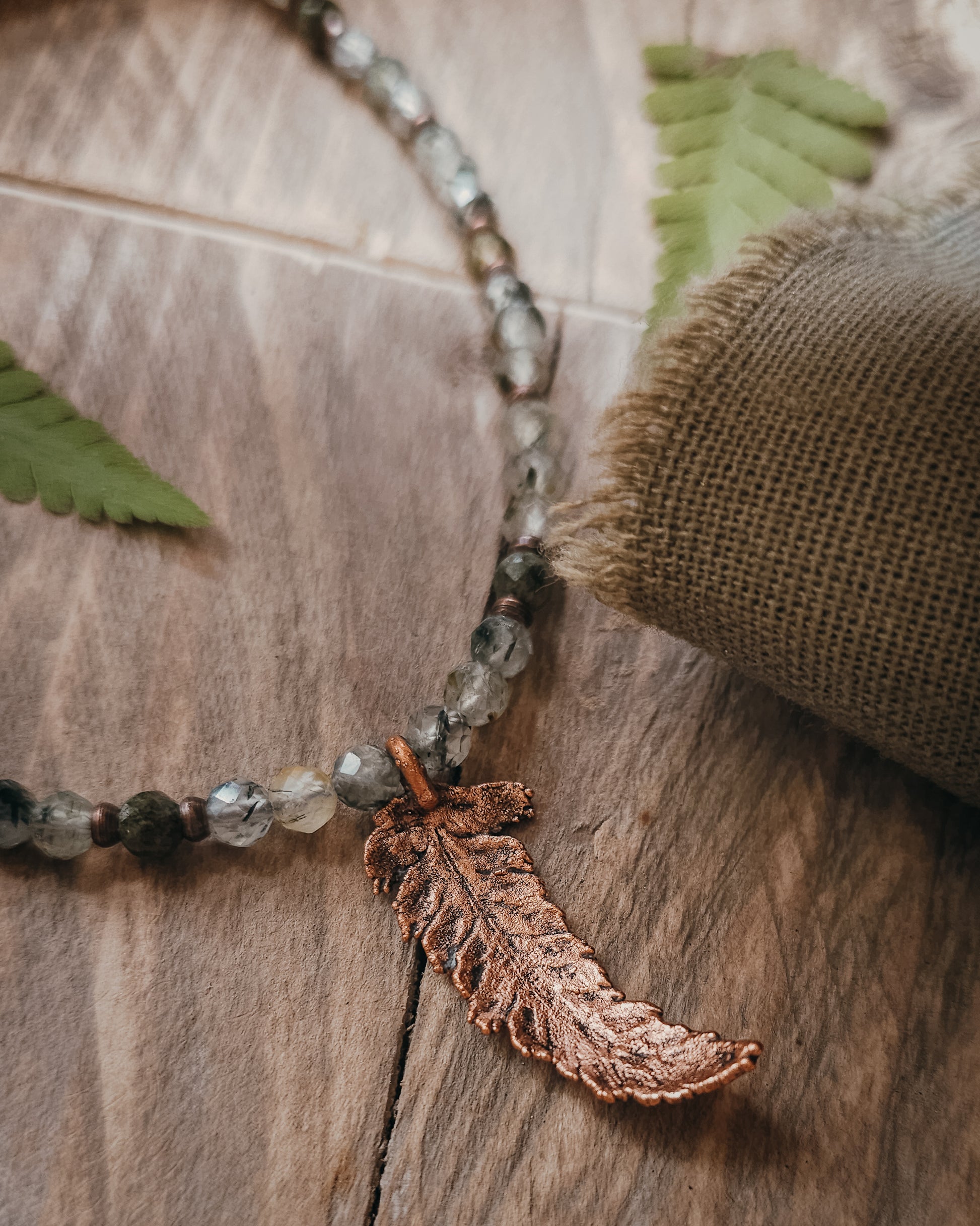 Close of copper electroformed fern leaf pendant and crystal Prehnite and Epidote beads necklace on a wooden surface with fern leaves