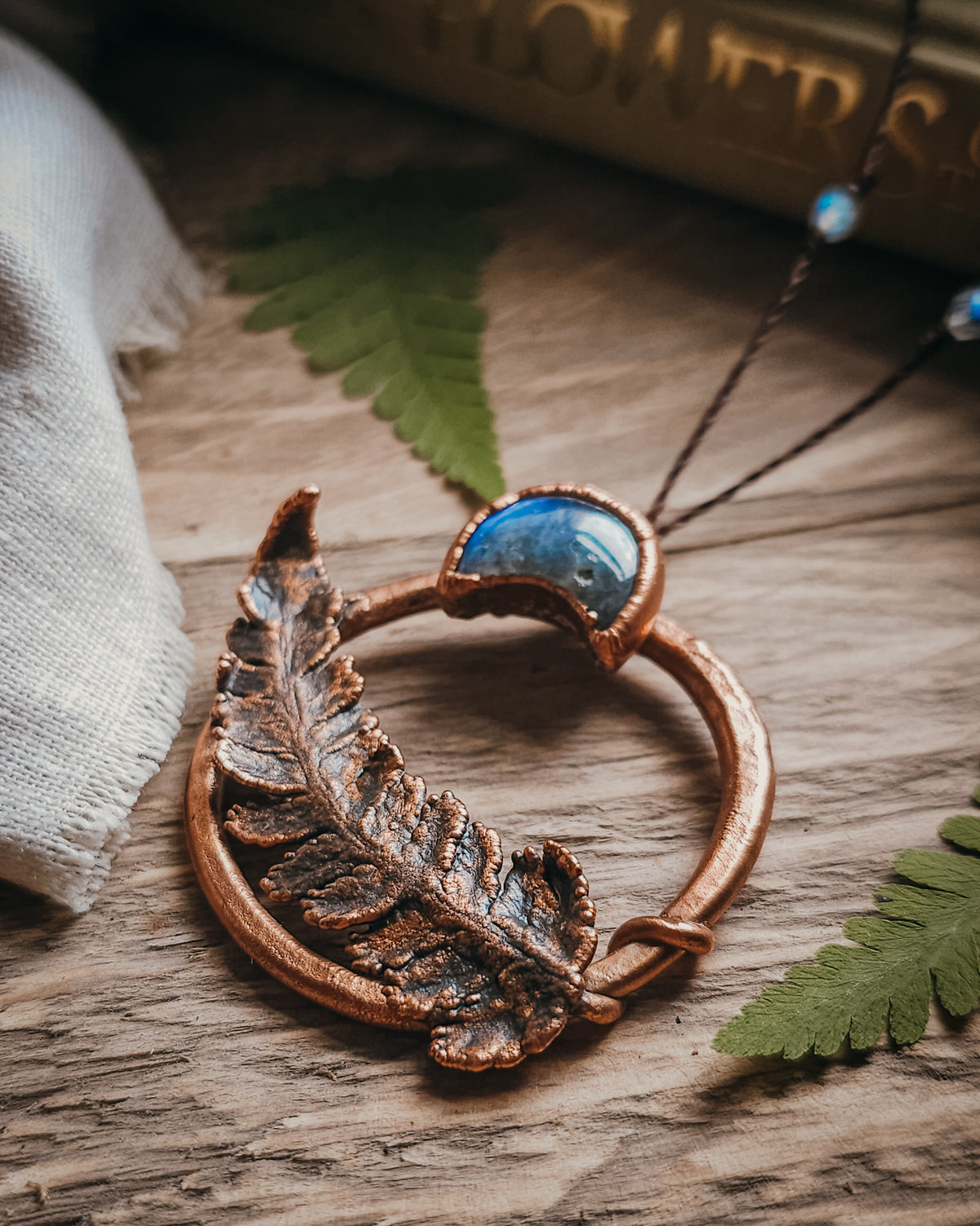 Copper electroformed fern pendant with a Labradorite crescent moon set within a circular copper frame, suspended from an adjustable cord with Labradorite and brass accent beads, handcrafted from a real fern gathered in Scotland and photographed on a rustic woodland-inspired background.