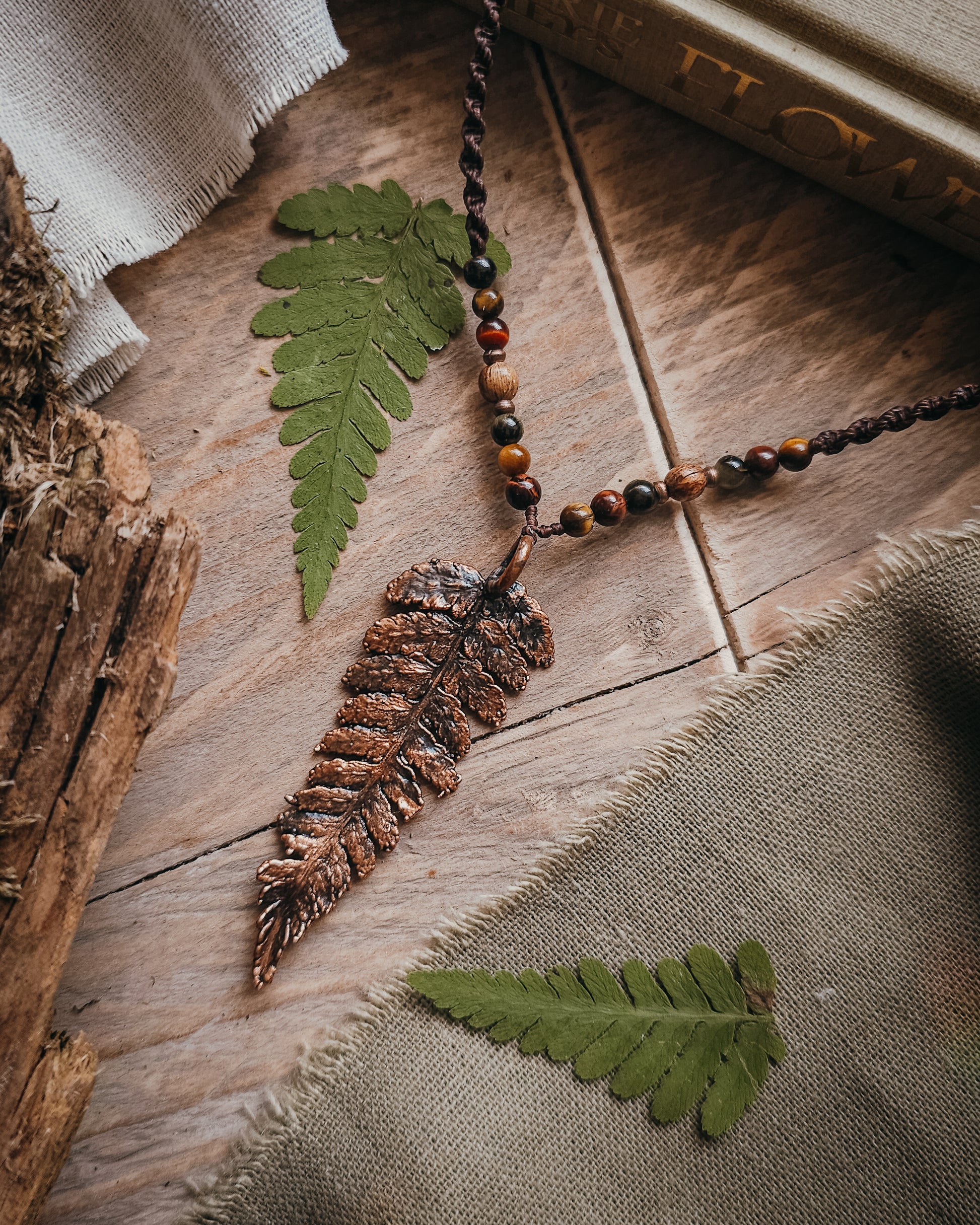 Necklace with  tiger's eye and wooden beads and a copper electroformed fern charm on a wooden surface with linen and fern leaf.