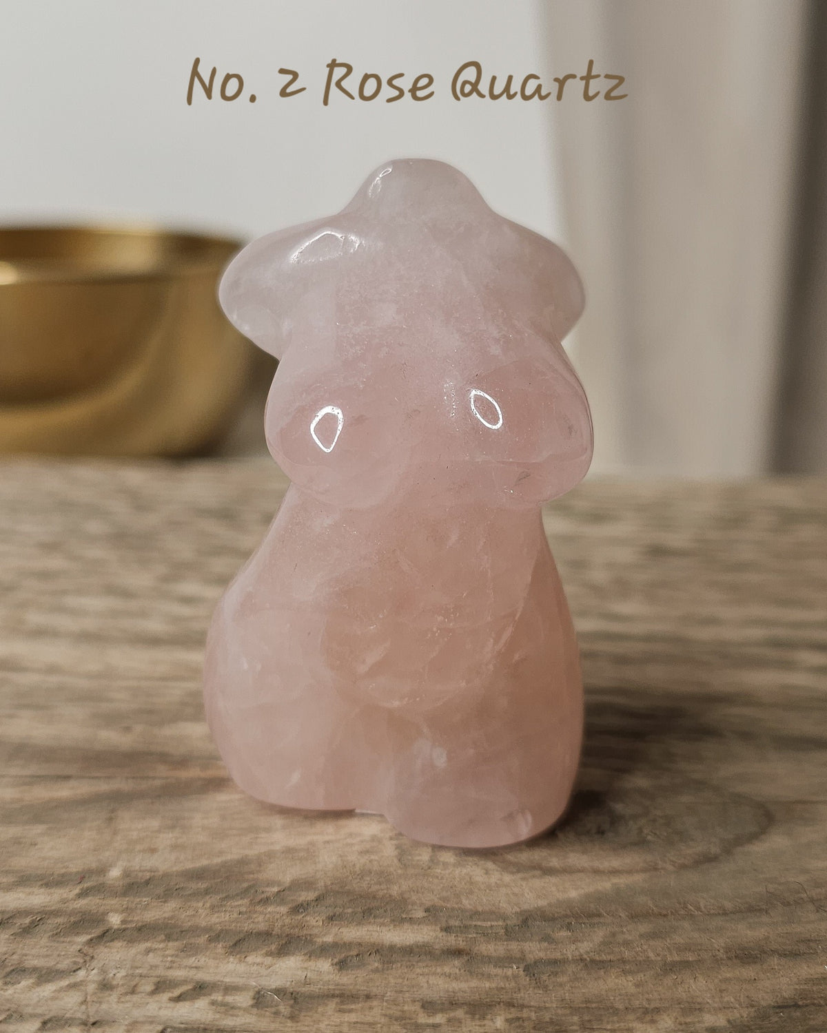 A small carved rose quartz figurine shaped like a female body, standing on a rustic wooden surface with a softly blurred indoor background.