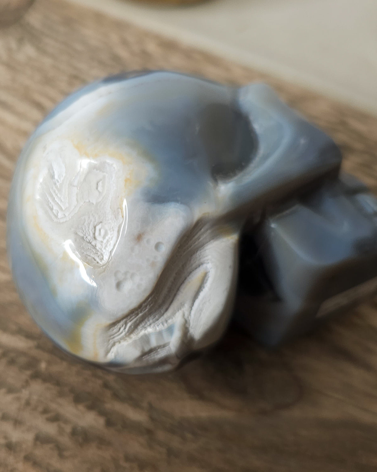 Blue Agate skull carving with layered pale blue and grey bands, a natural brown inclusion along the side and a sparkling druzy crystal cavity at the top.