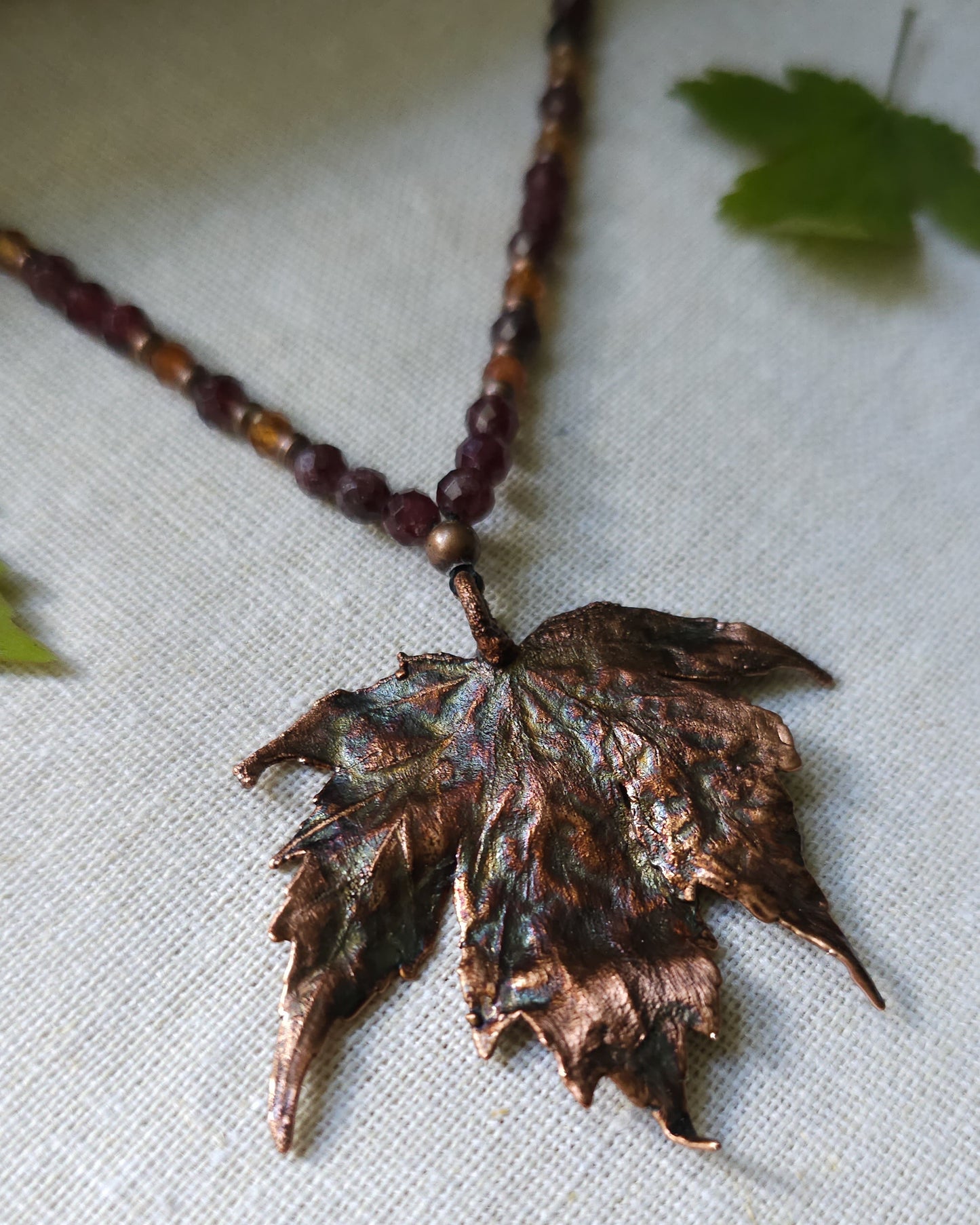 Handmade Copper electroformed Necklace with maple leaf pendant and Garnet crystal beads on a textured surface with leaves