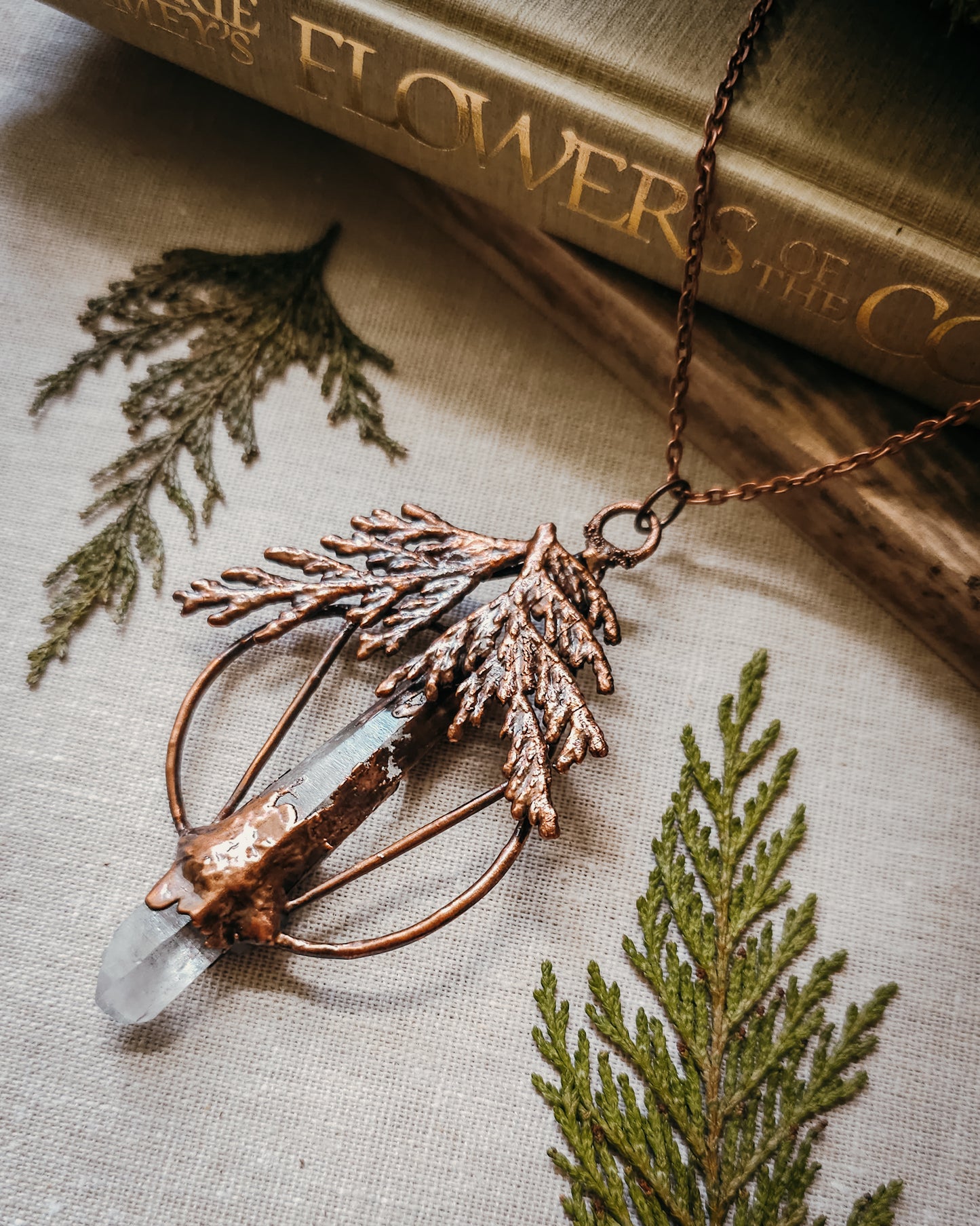 Handmade Copper electroformed Necklace with a cypress and clear Quartz crystal pendant on a textured fabric background