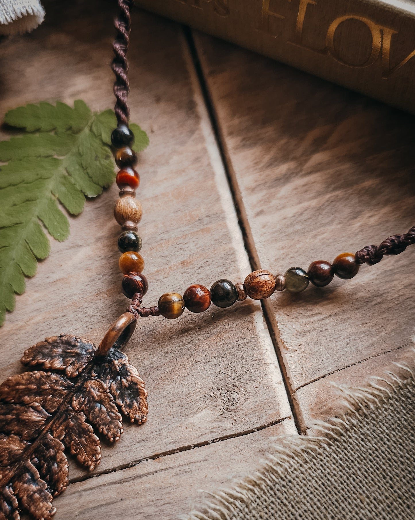 Close-up of Necklace with  tiger's eye and wooden beads and a copper electroformed fern charm on a wooden surface with linen and fern leaf.