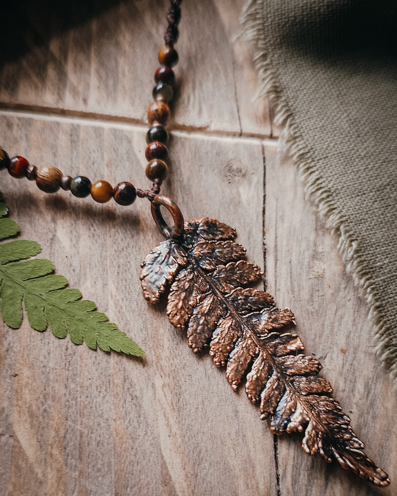 Close-up of Necklace with  tiger's eye and wooden beads and a copper electroformed fern charm on a wooden surface with linen and fern leaf.