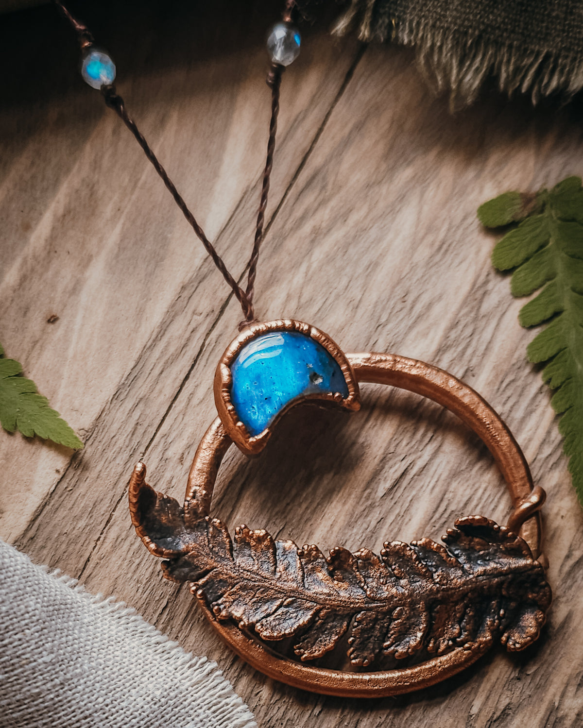 Copper electroformed fern pendant with a Labradorite crescent moon set within a circular copper frame, suspended from an adjustable cord with Labradorite and brass accent beads, handcrafted from a real fern gathered in Scotland and photographed on a rustic woodland-inspired background.