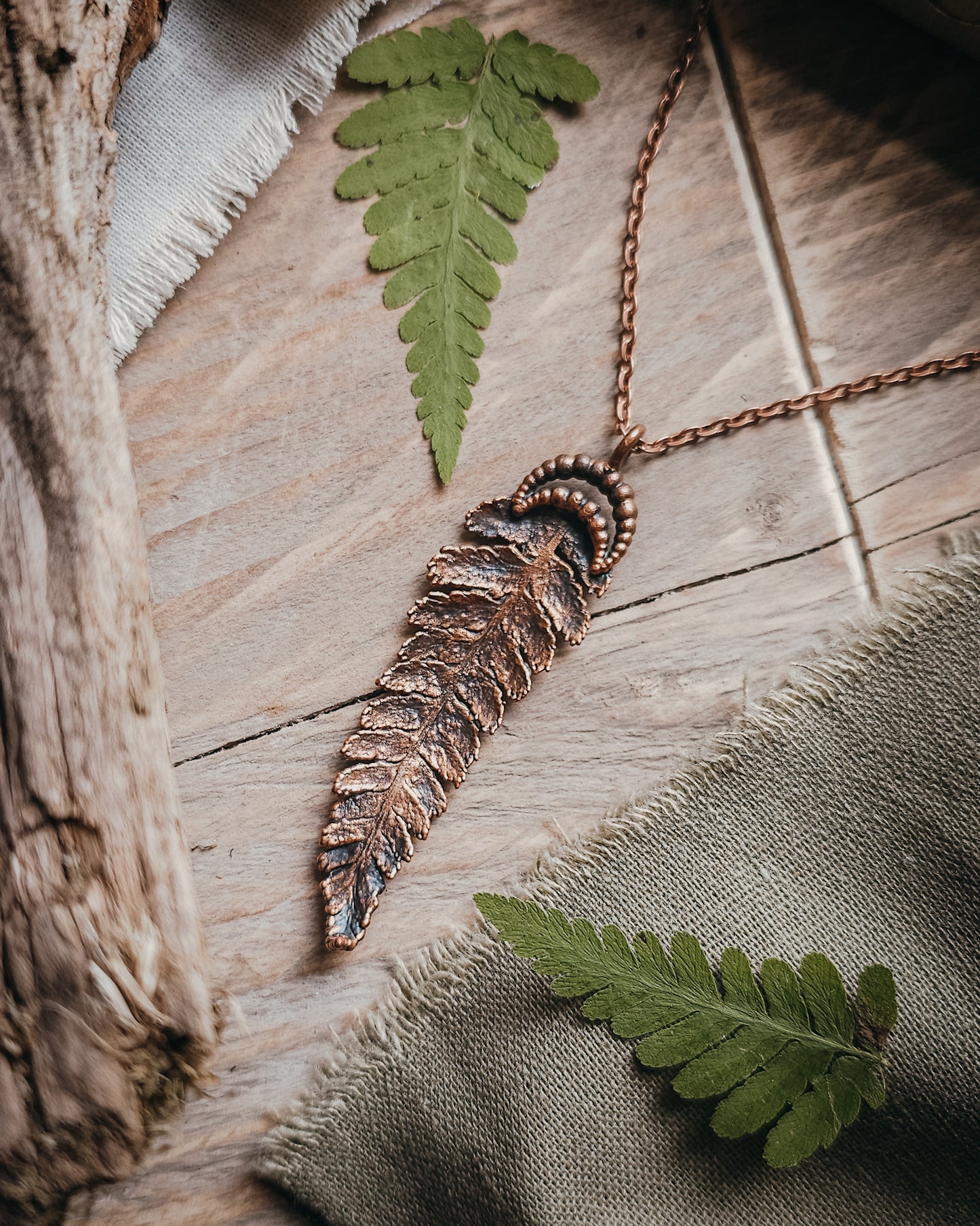 Copper electroformed fern pendant with crescent moon detail on a copper-coloured stainless steel chain, handcrafted from a real fern gathered in Scotland and photographed with fresh fern fronds on a rustic woodland-inspired background.