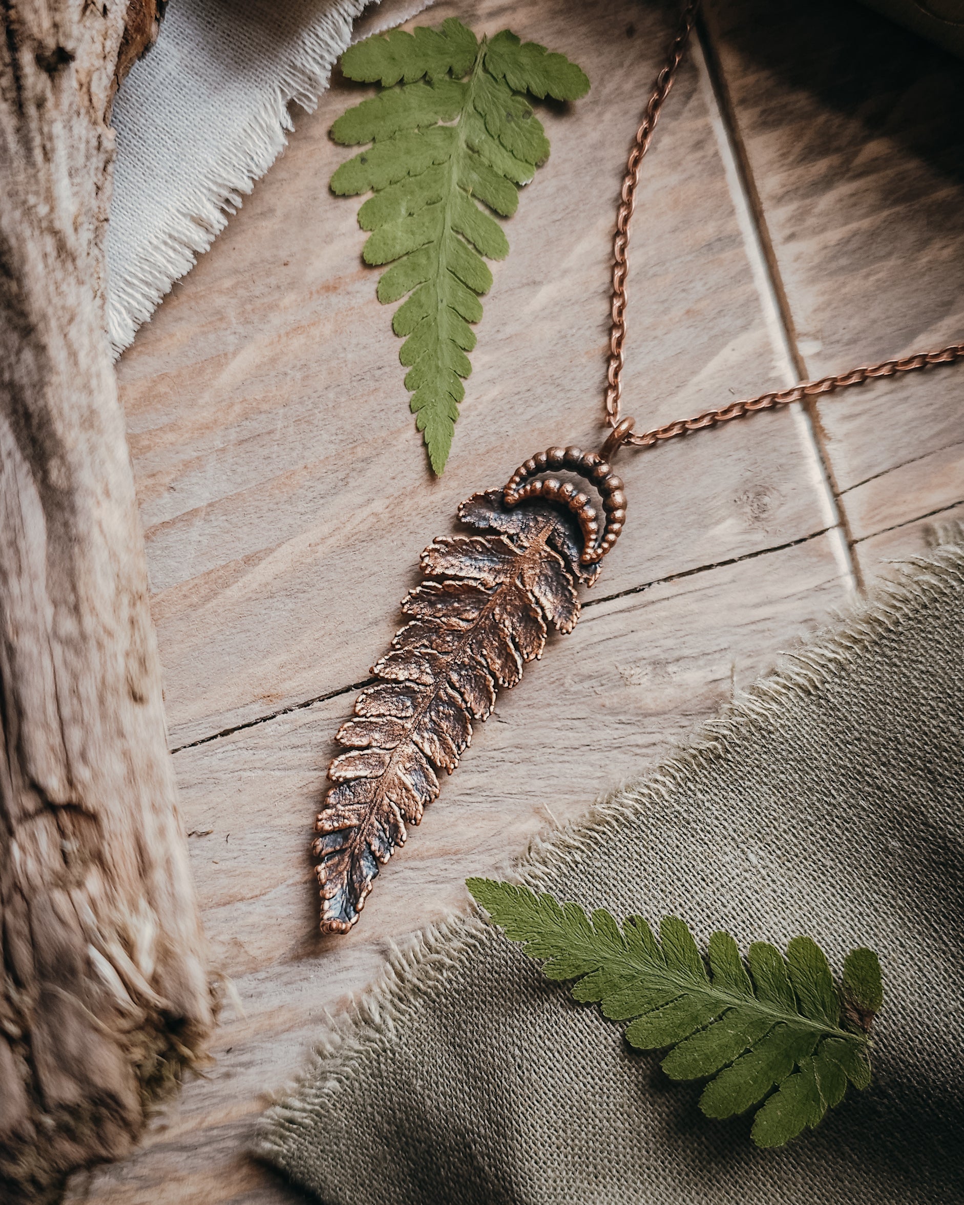 Copper electroformed fern pendant with crescent moon detail on a copper-coloured stainless steel chain, handcrafted from a real fern gathered in Scotland and photographed with fresh fern fronds on a rustic woodland-inspired background.