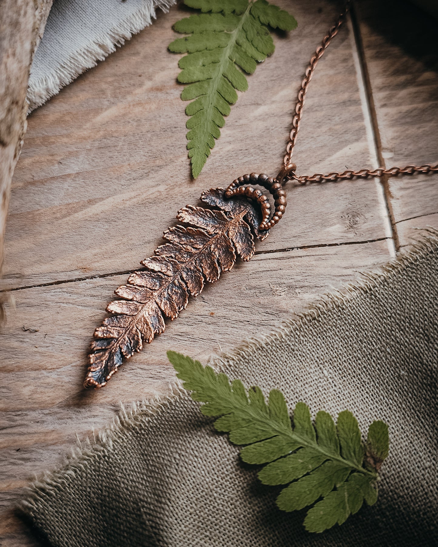 Copper electroformed fern pendant with crescent moon detail on a copper-coloured stainless steel chain, handcrafted from a real fern gathered in Scotland and photographed with fresh fern fronds on a rustic woodland-inspired background.