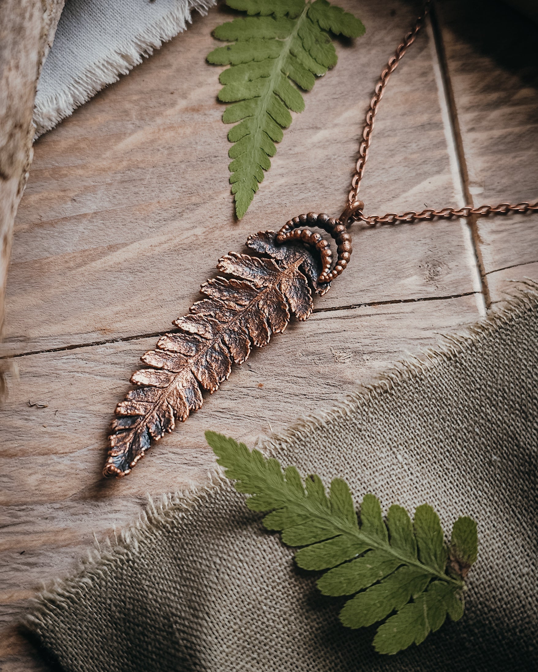 Copper electroformed fern pendant with crescent moon detail on a copper-coloured stainless steel chain, handcrafted from a real fern gathered in Scotland and photographed with fresh fern fronds on a rustic woodland-inspired background.