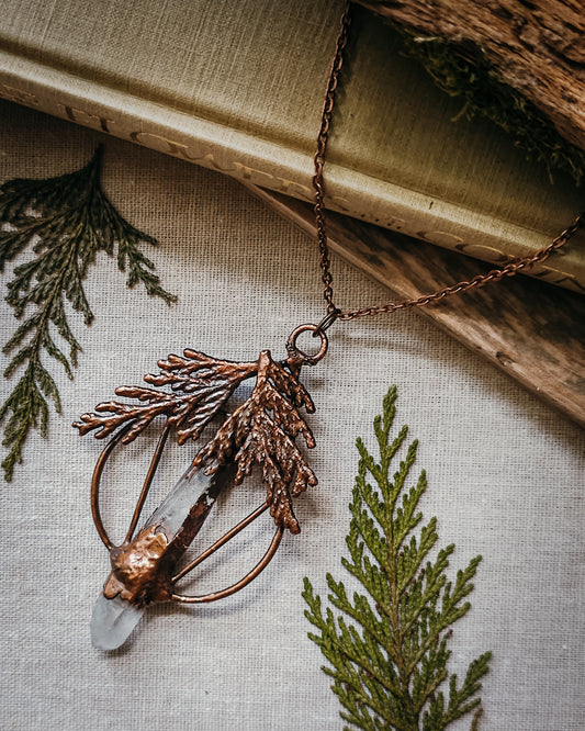Handmade Copper electroformed Necklace with a cypress and clear Quartz crystal pendant on a textured fabric background