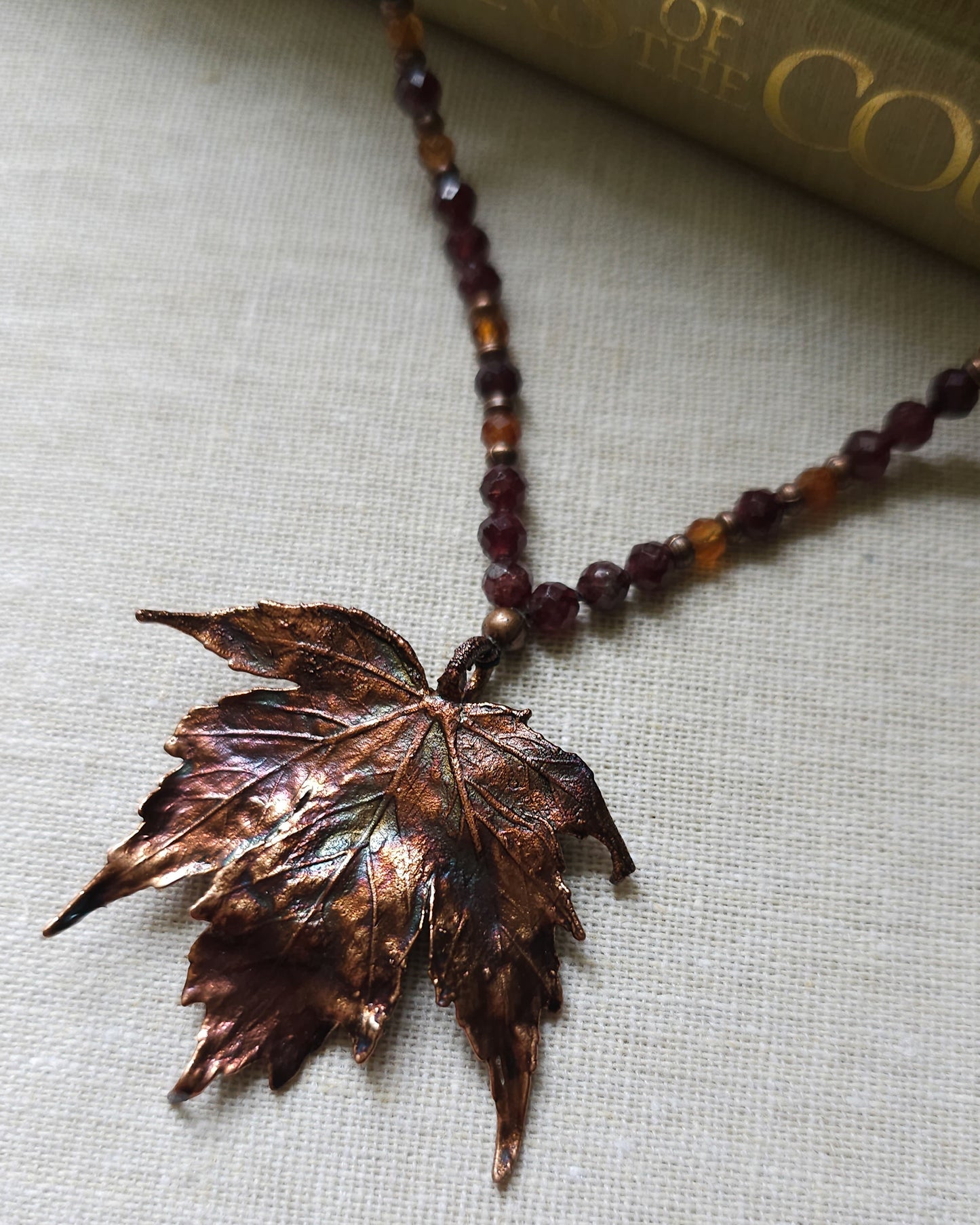 Handmade Copper electroformed Necklace with maple leaf pendant and Garnet crystal beads on a textured surface with leaves