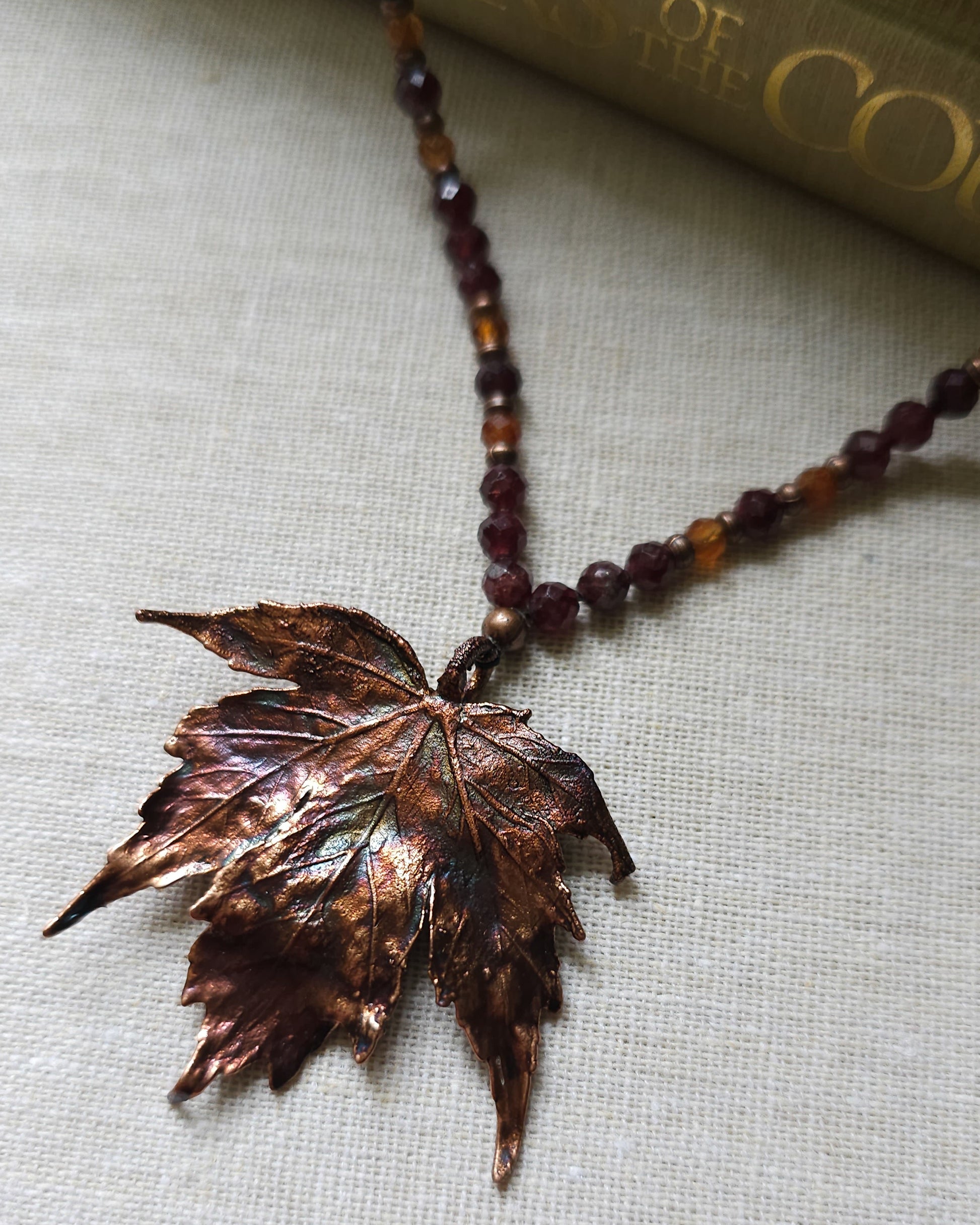 Handmade Copper electroformed Necklace with maple leaf pendant and Garnet crystal beads on a textured surface with leaves