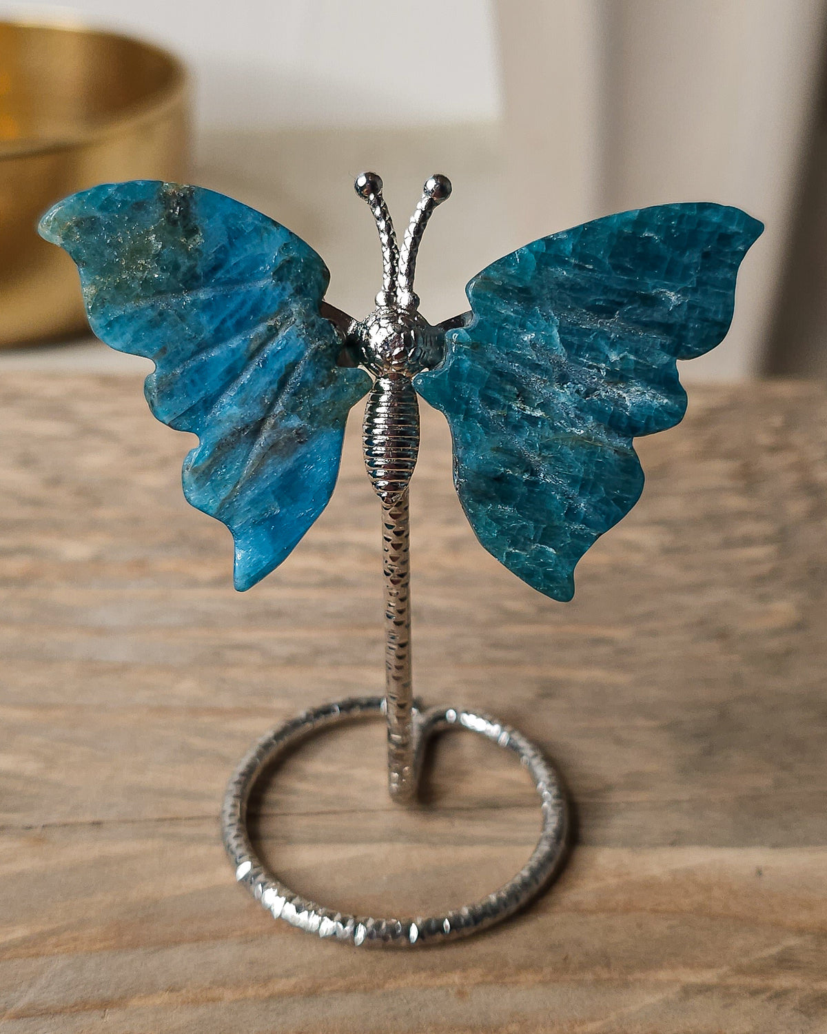 Blue Apatite butterfly carving on a decorative silver-tone metal stand, featuring rich ocean-blue and teal colouring with natural mineral patterns and carved wing details.
