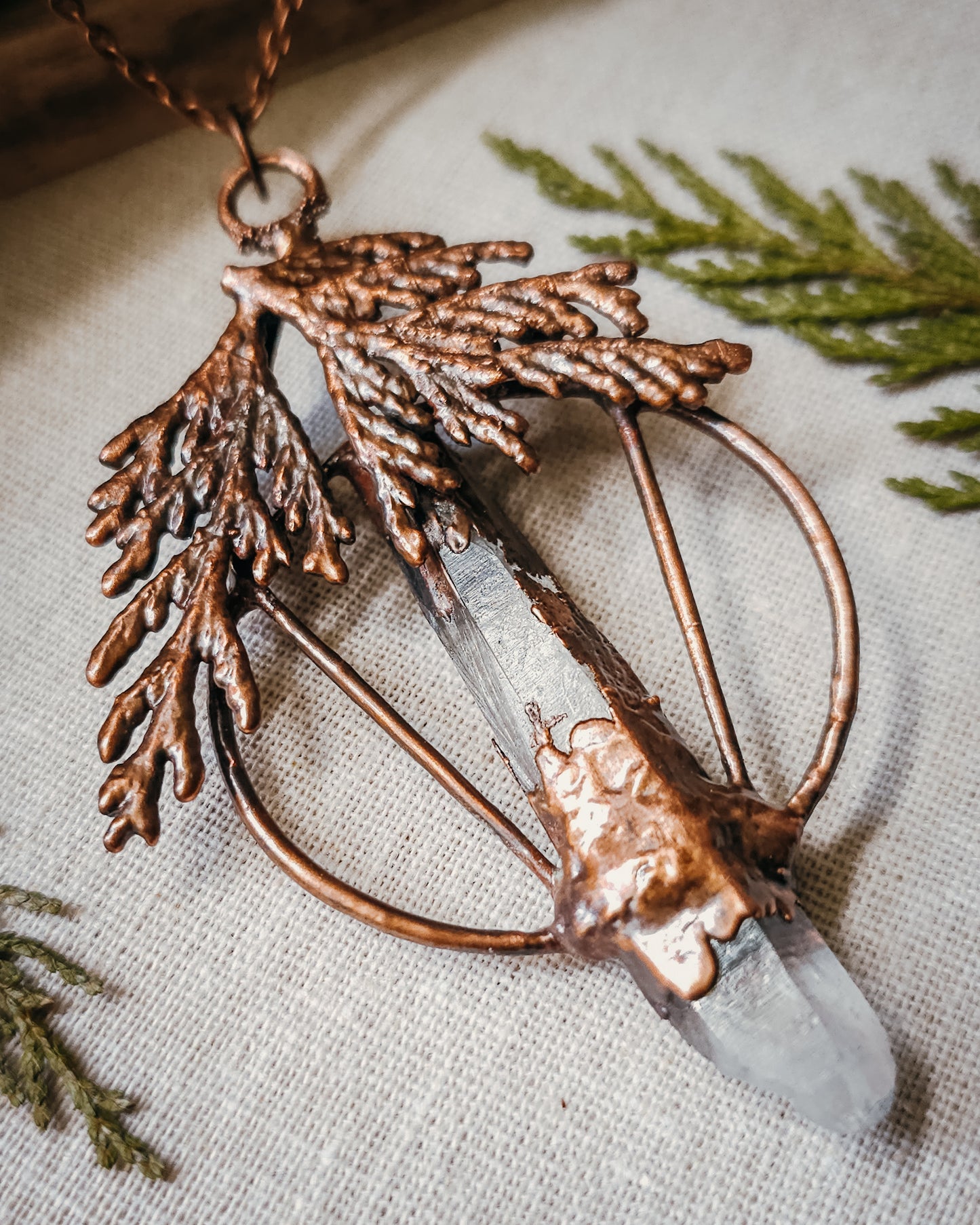 Handmade Copper electroformed Necklace with a cypress and clear Quartz crystal pendant on a textured fabric background