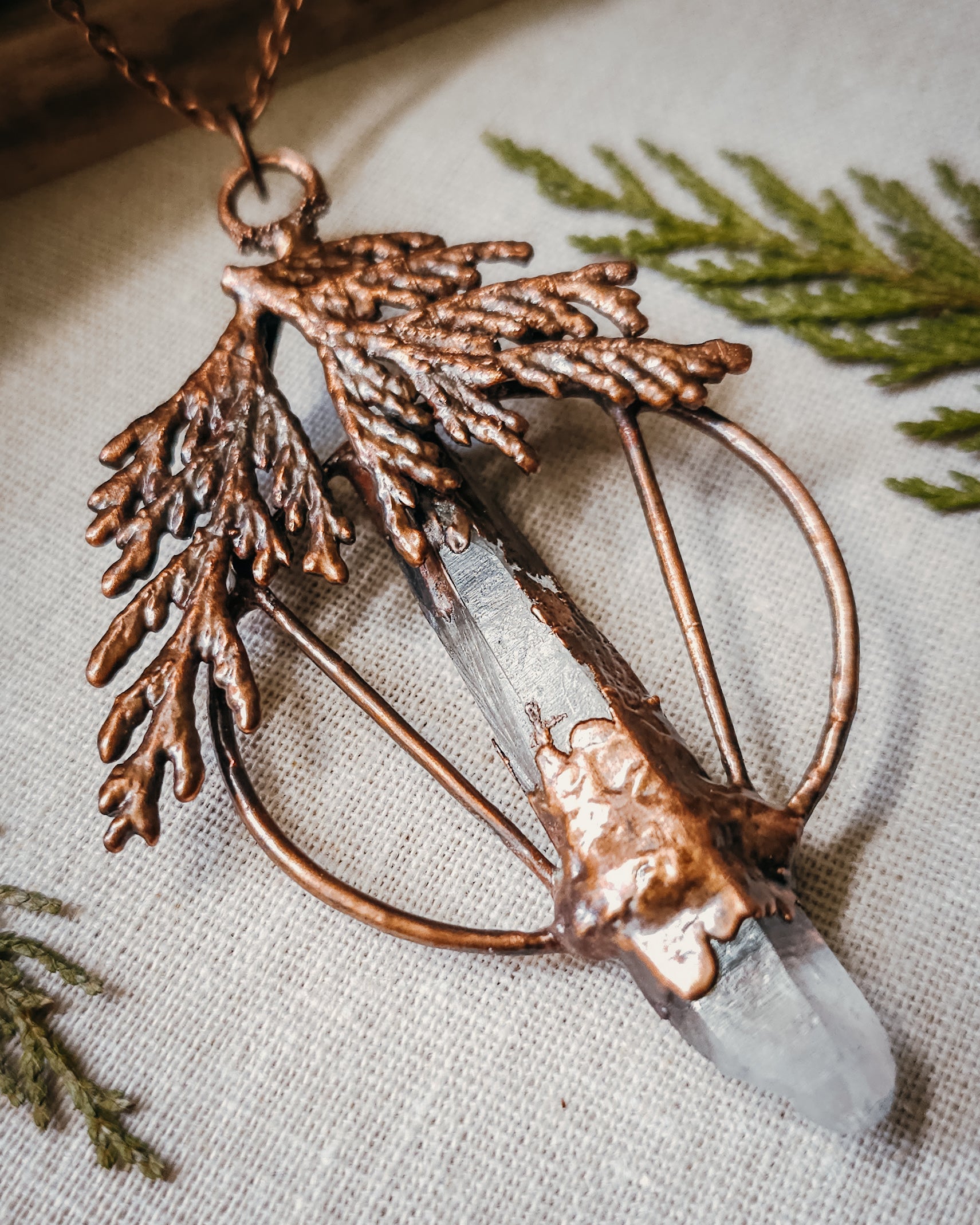 Handmade Copper electroformed Necklace with a cypress and clear Quartz crystal pendant on a textured fabric background
