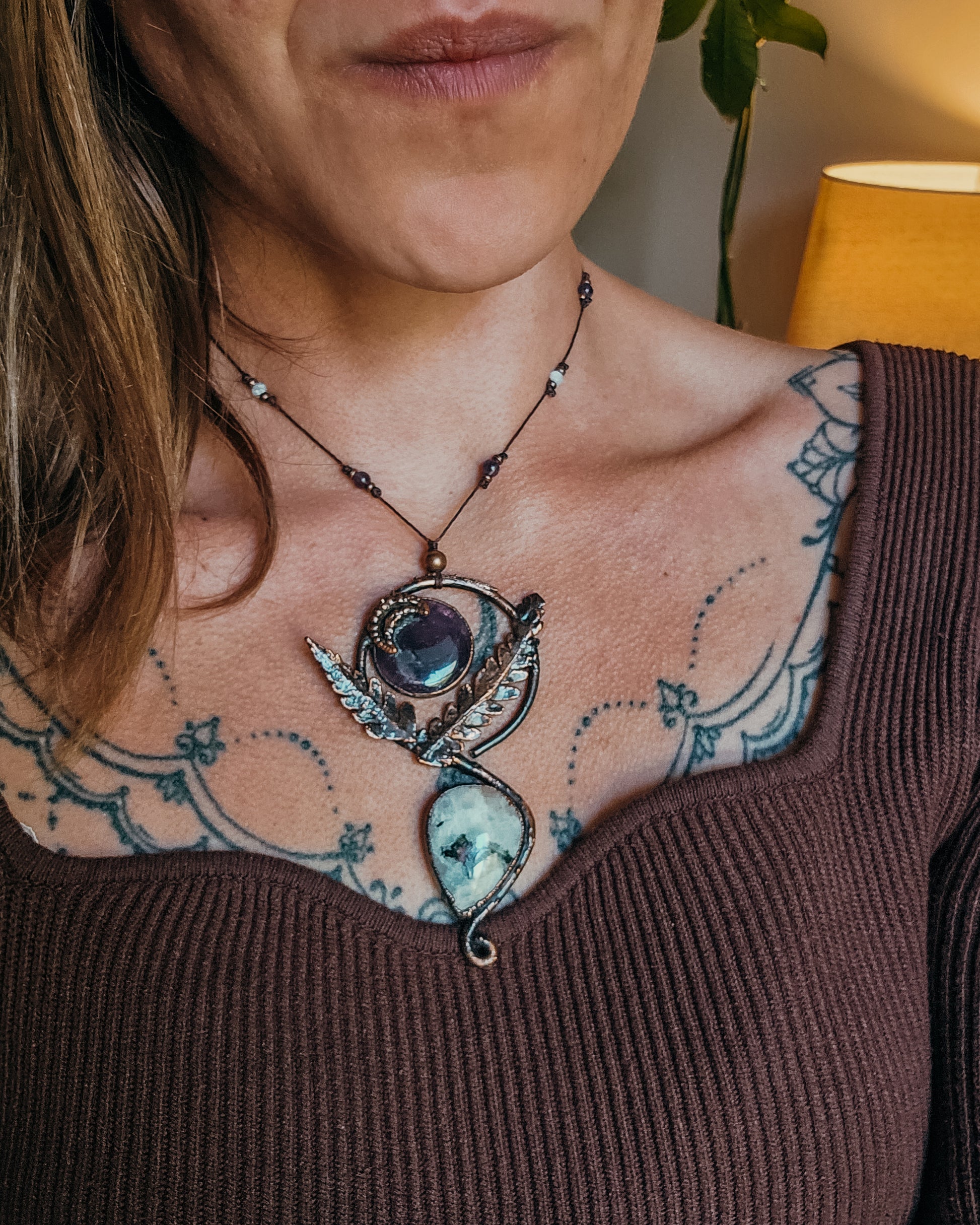 Person wearing a Copper electroformed Ornate pendant with amehtyst, Moonstone and fern leaves on a wooden surface with leaves with a brown top and visible tattoos.