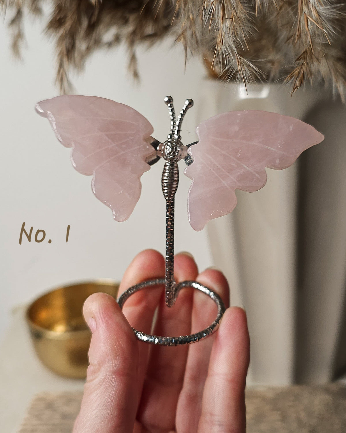 Polished rose quartz crystal butterfly carving with soft pink wings on a silver-tone display stand. 

